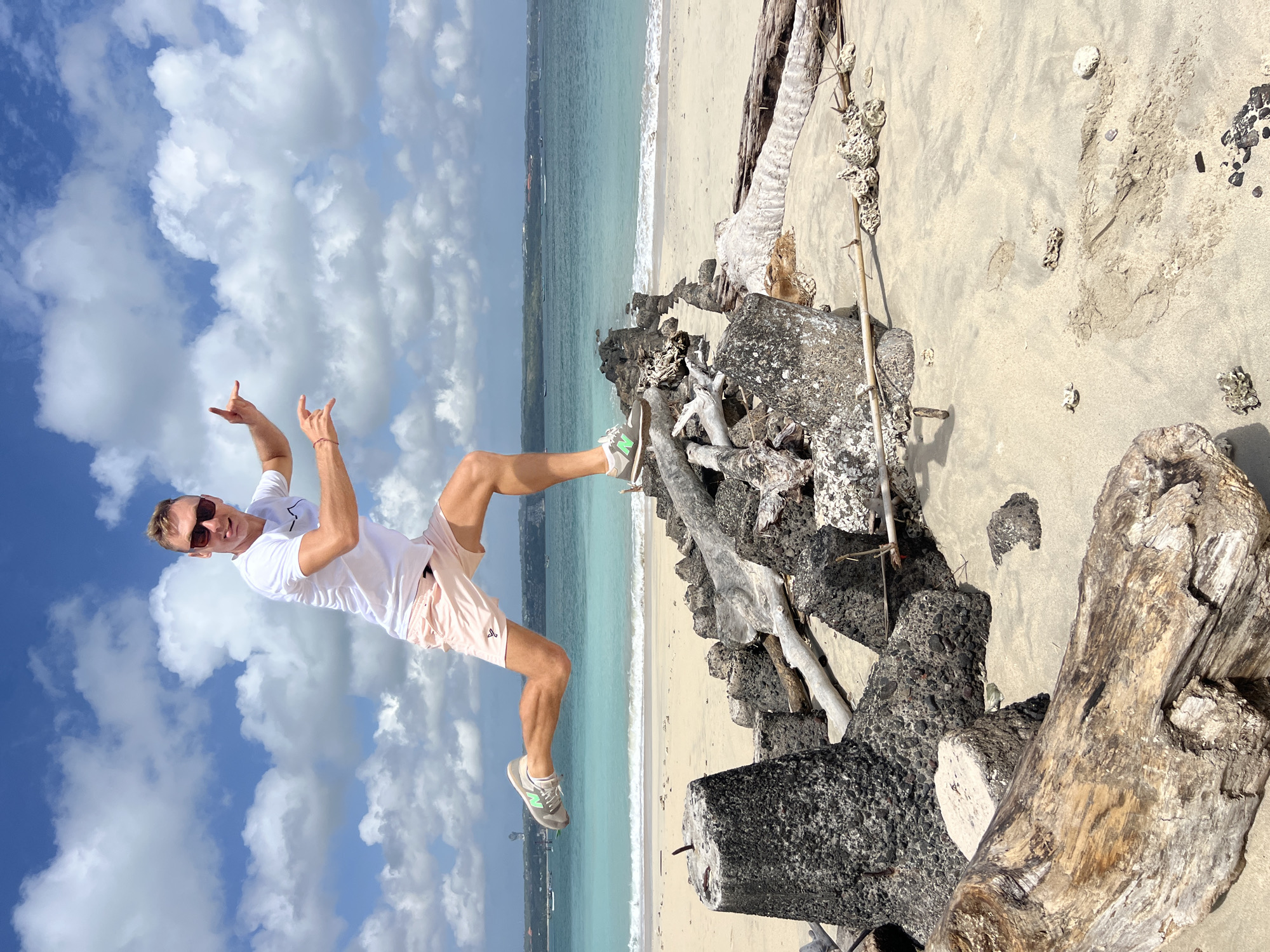 A man is mid-air, jumping over a pile of rocks and driftwood on a sandy beach. He is wearing a white t-shirt, light pink shorts, sunglasses, and athletic shoes. His left leg is extended forward, and his right leg is bent behind him. His arms are outstretched, with his right hand making a "rock on" gesture. The beach has a turquoise ocean with gentle waves in the background, and a clear blue sky with white clouds above. The location is Denpasar, Indonesia.