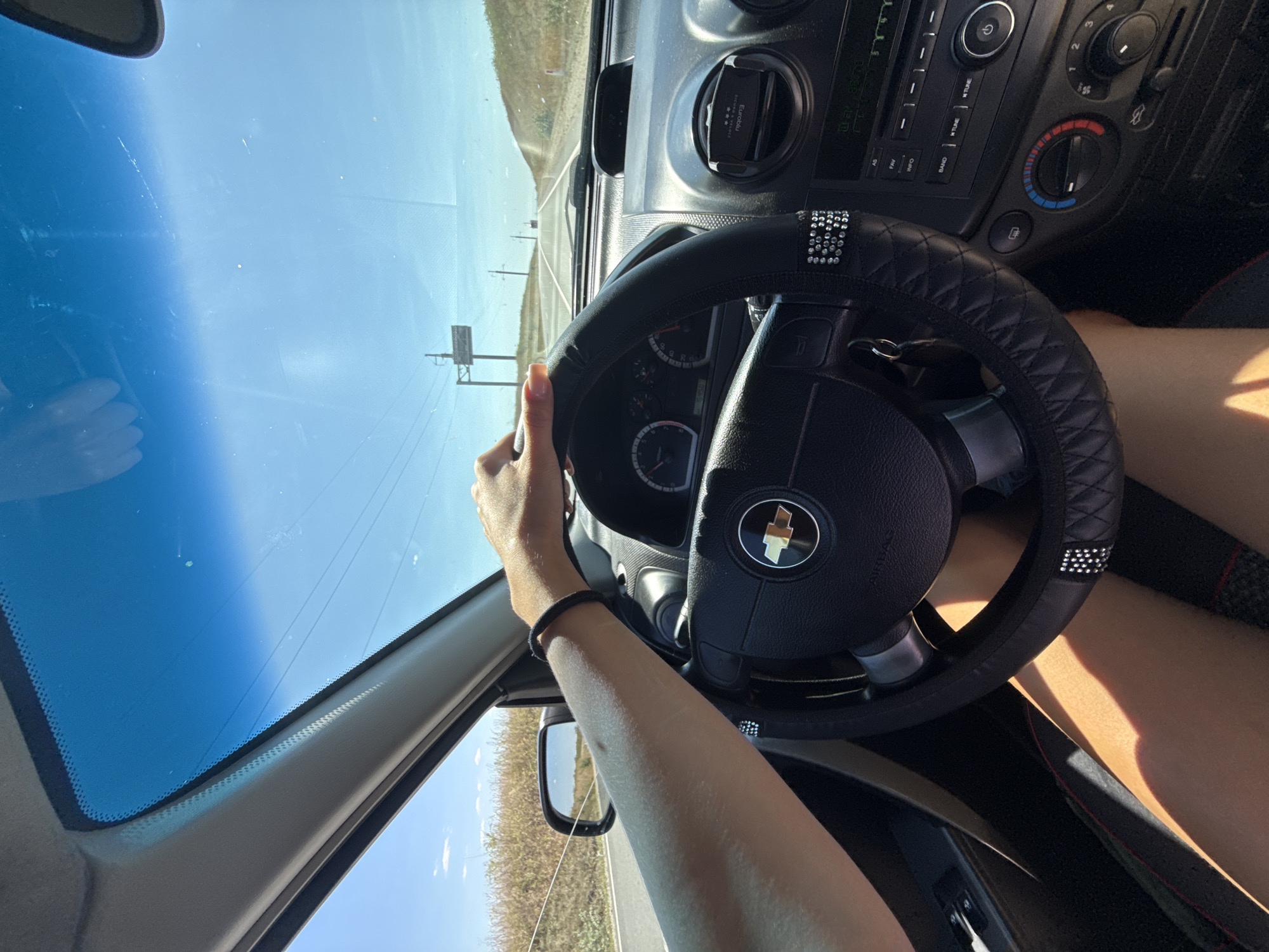 This is a first-person perspective shot from inside a car driving on a sunny day. The driver's hands are on the steering wheel. The dashboard and the road ahead are visible. The car appears to be on a highway or a country road, with dry vegetation on the sides. The sky is bright blue. The image appears to be taken in Cioc-Maidan, Moldova, a rural area known for its agricultural landscape and roads.