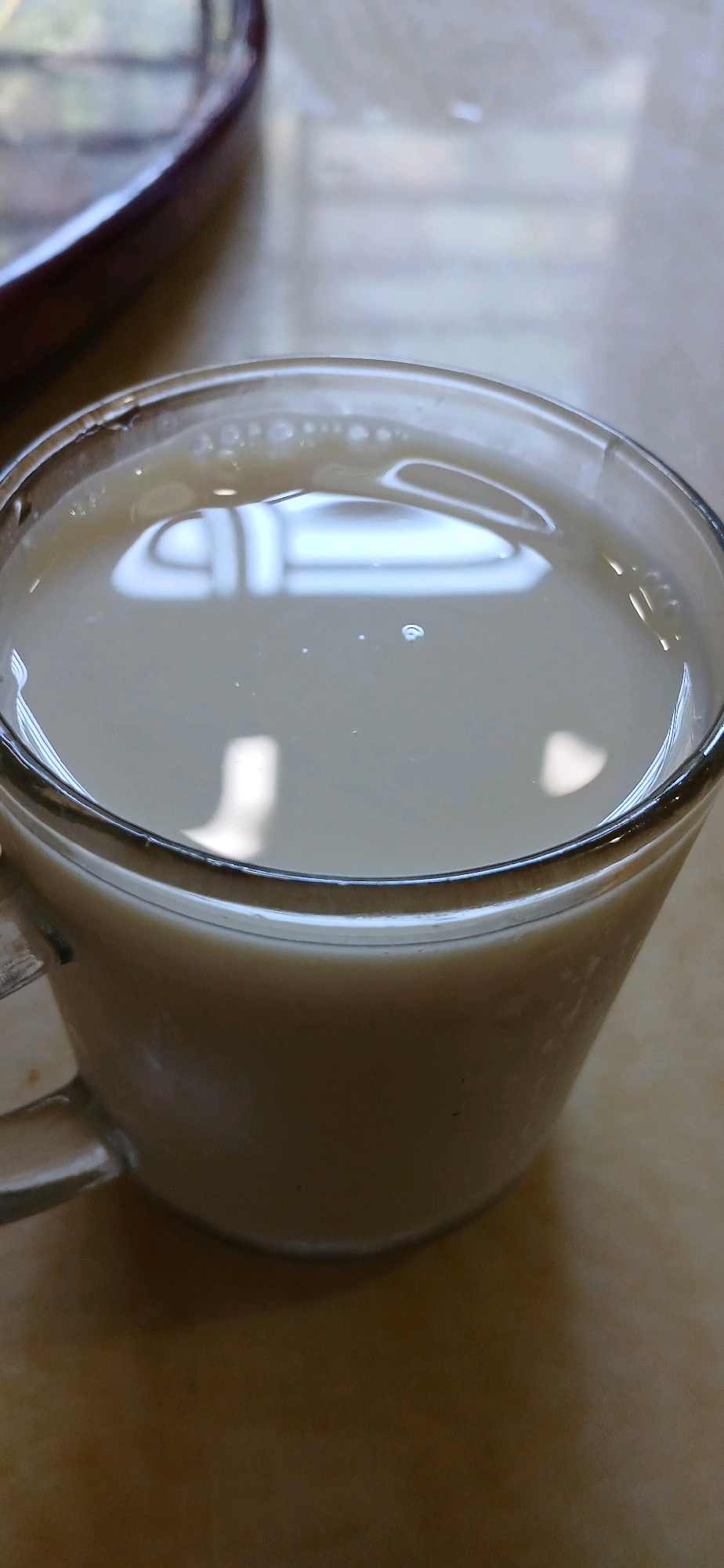 A close-up shot shows a glass mug filled with a light-colored liquid, possibly milk or tea, with small bubbles visible on the surface. The mug is clear glass with a handle on the left side. Reflections of light and what appear to be window panes are visible on the liquid's surface. The mug sits on a textured surface that appears to be a table, with a blurred dark object in the upper left corner. The overall scene suggests a casual setting, possibly a home or cafe. The location is identified as Tanjung Pinang, Indonesia, though there are no specific visual cues in the image to confirm this.