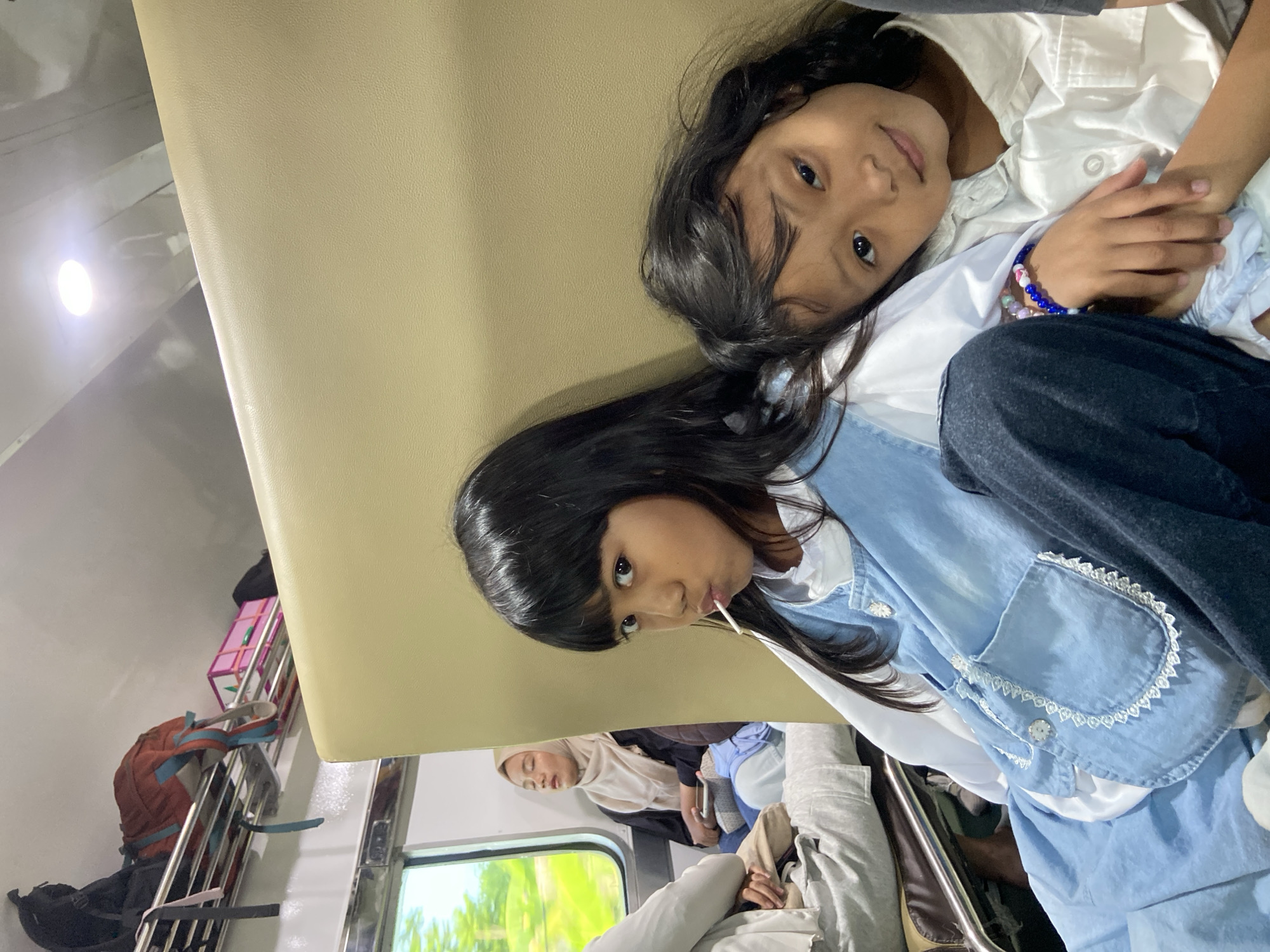 Two young girls with long black hair are lying down in what appears to be the overhead luggage area of a train. One girl is wearing a white collared shirt and beaded bracelet, while the other is wearing a light blue denim jacket and holding a lollipop in her mouth. Other passengers can be seen sitting in the seats below. Luggage, including a backpack and a pink box, is stored on the overhead rack. The scene is taking place on a train, presumably traveling through or near Dandang Gendis, Indonesia.