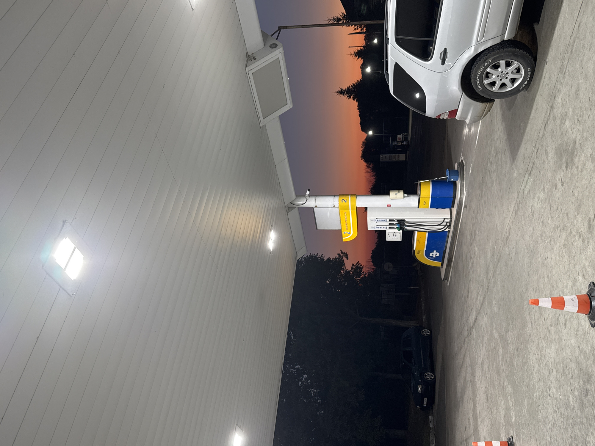 A gas station canopy in Sredets, Bulgaria, is illuminated by overhead lights. The sky displays a gradient of sunset colors, from deep orange near the horizon to purple and dark blue above. Two cars are present: a dark sedan parked on the left side of the frame, and the rear of a silver SUV on the right. A gas pump with yellow and blue branding stands in the center. Several orange and white traffic cones are placed on the concrete ground. Trees and some streetlights are visible in the background, silhouetted against the twilight sky.