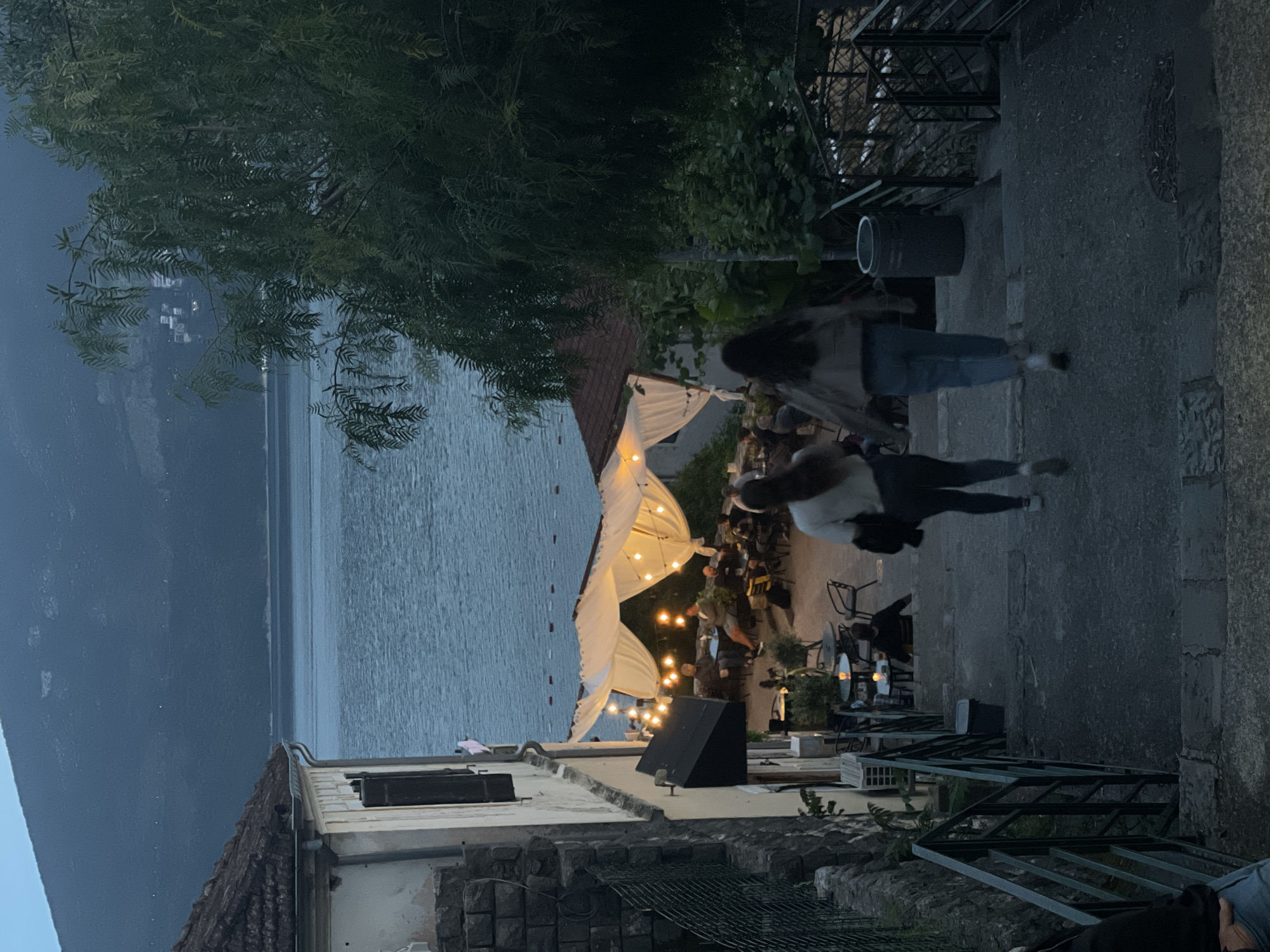 The image shows an outdoor restaurant scene in Herceg Novi, Montenegro, overlooking a body of water.
**Environment:** The setting is a hillside location within the city. Buildings with stone walls and tiled roofs are visible in the foreground. The landscape extends towards a large body of water (likely the Bay of Kotor) with mountains visible in the distance. The lighting suggests it's dusk or evening.
**Objects:** The outdoor restaurant is covered by a white canopy and illuminated with string lights. There are tables and chairs arranged for dining. A black speaker is visible on a rooftop. Stairways with railings lead up and down the hillside.
**People:** Two people are walking up a set of stone steps. A group of people are seated at the restaurant tables, seemingly dining or conversing.
**Actions/Interactions:** The people on the stairs appear to be in motion, heading uphill. The restaurant patrons are engaged in activities associated with dining.