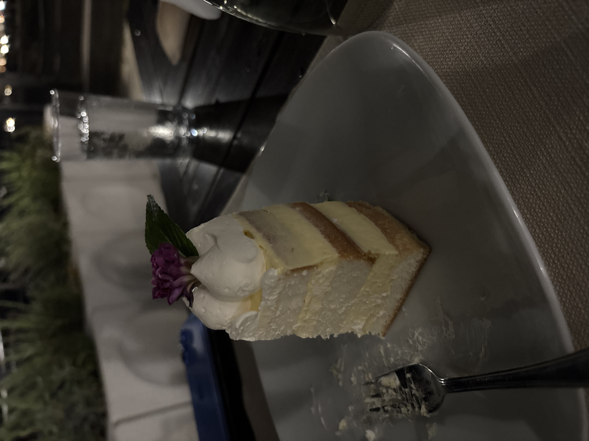 A slice of layered cake with white frosting and a purple flower on top sits on a white plate. A fork with some frosting on it is to the left of the cake. In the background, a glass of water is on a dark wooden table. The scene appears to be outdoors, possibly at a restaurant or cafe, with greenery and a blurry background. The lighting suggests it is nighttime. The location is Varna, Bulgaria.