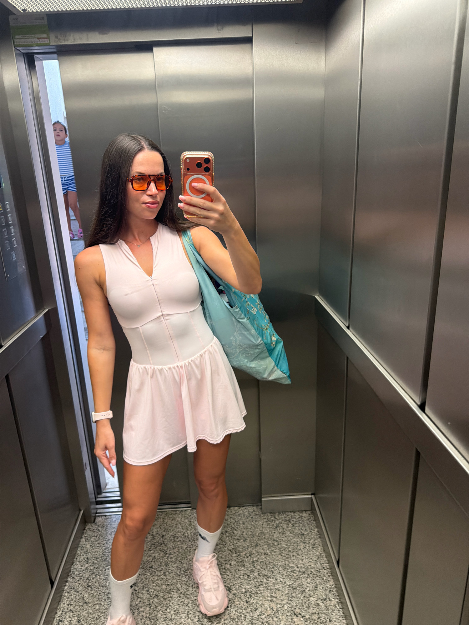 An individual is taking a selfie in an elevator. The person is wearing an athletic-style pink dress with a zippered neckline, white socks, and light pink athletic shoes. They also have orange-tinted sunglasses and a light blue tote bag with a nature-inspired print. A child in a striped shirt and shorts is visible in the background, standing near the elevator doors. The elevator is metallic with textured walls and a grey speckled floor. The overall scene suggests an urban environment in Salou, Spain, possibly at a resort or hotel given the presence of a child and the casual attire.
