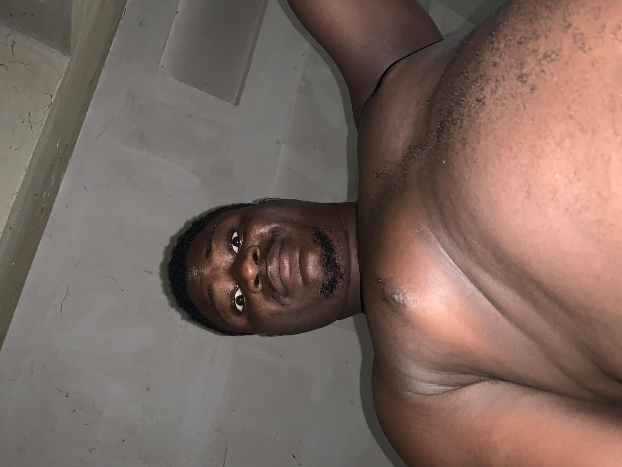 The image is a close-up, low-angle shot of a Black man's face and upper body, taken in Akwanga, Nigeria. The man is looking directly at the camera with his head tilted. His expression appears neutral, with his eyes open and looking forward. He has short, dark hair and a light beard. The background consists of a grey, textured wall, possibly a building interior. The lighting is dim, suggesting it might be nighttime or indoors with limited light. The image is rotated 90 degrees counter-clockwise, so the man appears to be lying on his side. There is no visible text in the image. The focus is on the man's features and skin texture.