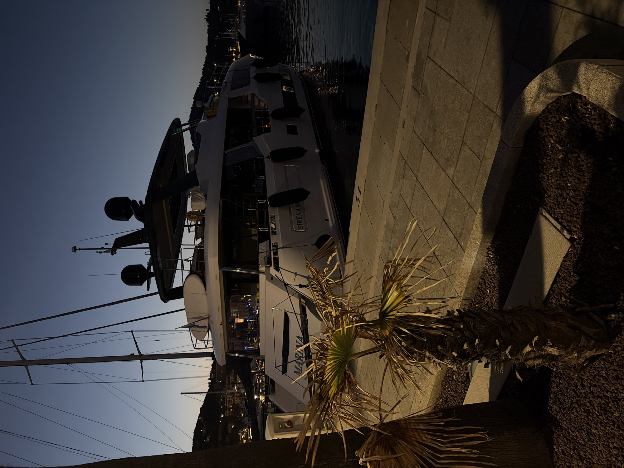 A modern, white yacht named "MARINA" is moored at night in the Port of Imperia, Italy. The yacht, which also has "SIRENA" visible on its hull, is illuminated by internal and external lights, casting reflections on the dark water. Several black fenders are attached to the yacht's side. A paved walkway, likely part of the marina, runs along the edge of the water, with a low concrete border containing dark gravel and two small palm trees. One palm tree in the foreground has dry, brown fronds, while the other is partially visible to its left. In the background, other boats and masts are visible, along with lights from the city lining the hillsides. The sky is a deep twilight blue. The scene depicts a still, quiet evening at the harbor.