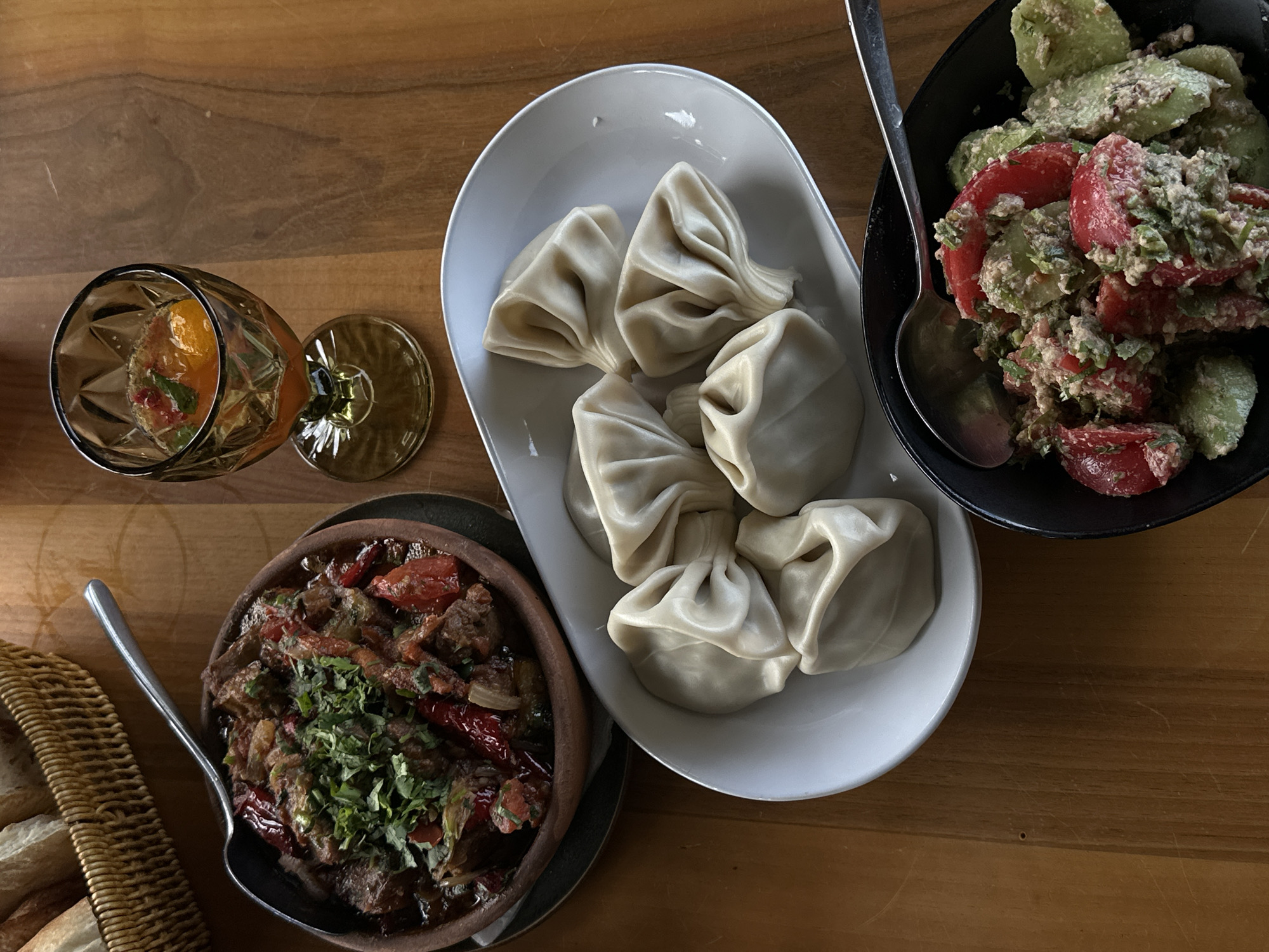 The image shows a wooden table set with a selection of Georgian dishes, likely in the city of Marneuli, Georgia.
On the table are:
* A white oval dish containing six khinkali, which are Georgian dumplings.
* A dark brown ceramic bowl filled with a stew-like dish. The stew contains pieces of meat, tomatoes, and red peppers, garnished with chopped herbs. A spoon is placed next to the bowl.
* A black bowl filled with a salad made from sliced tomatoes and cucumbers, tossed in a creamy sauce and garnished with herbs. A spoon is inside the bowl.
* A glass with a drink that contains sliced oranges.
* A wicker bread basket with slices of white bread.
The dishes and utensils are arranged in a manner typical of a shared meal. The scene suggests a dining experience centered around Georgian cuisine.