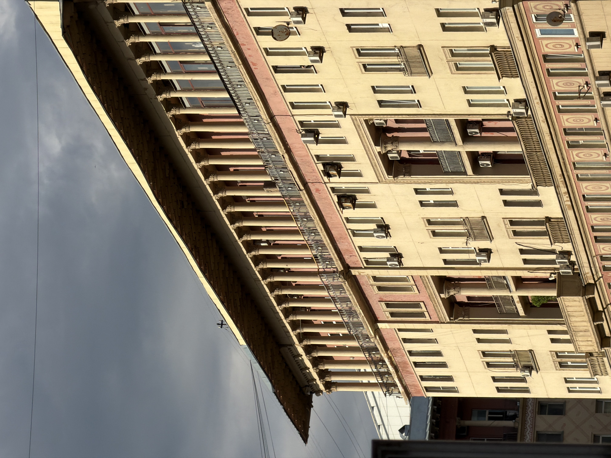 The image shows a fragment of a historic building in Moscow, Russia. The building is characterized by its yellow facade, numerous windows, balconies, and decorative elements such as columns and moldings. Air conditioning units are visible on the exterior walls, indicating modern adaptations to the structure. The sky is overcast, and a portion of a nearby building is visible in the lower right corner. The architectural style suggests a building constructed in the Stalinist Empire style, a common architectural style in Moscow during the mid-20th century.