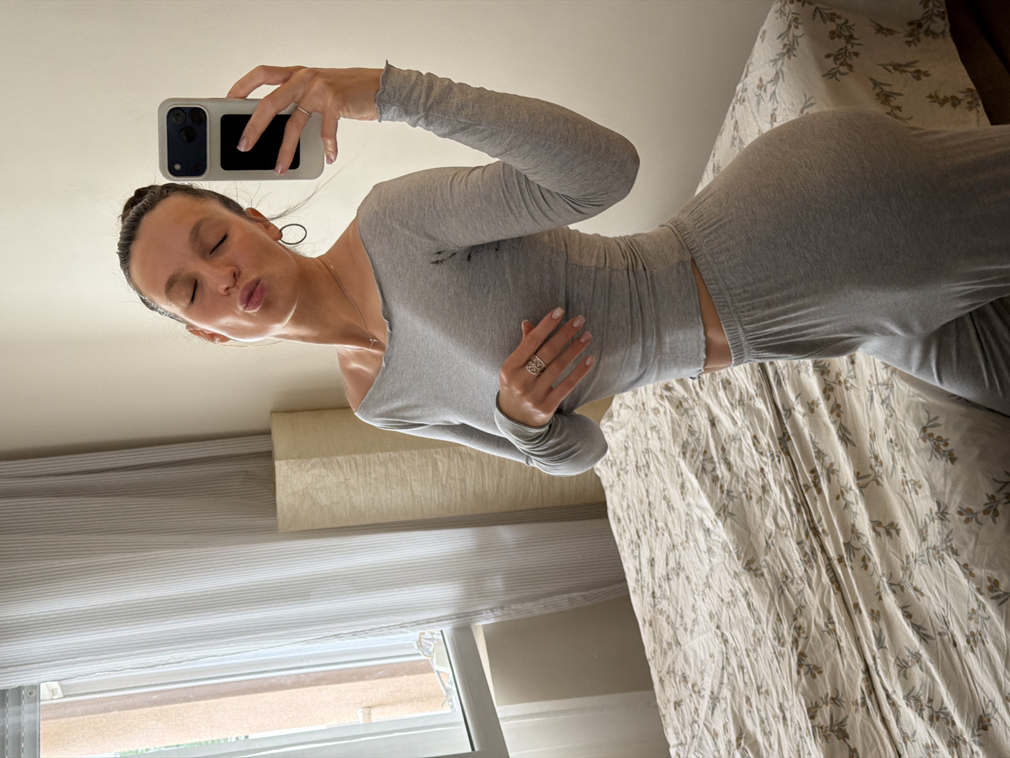 A young woman is captured in a mirror selfie, posing playfully with her eyes closed and lips pursed in a kissy face. She holds a light-colored smartphone with a multi-lens camera in her left hand, which also features a silver ring on her ring finger. She wears a form-fitting, light grey loungewear set, consisting of a long-sleeved crop top with ruffled cuffs and matching high-waisted pants that accentuate her figure. A delicate silver necklace with a small pendant and hoop earrings are visible. Another silver ring adorns her right hand's middle finger.
The reflection in the mirror reveals an indoor setting, likely a bedroom in Budva, Montenegro, during daylight hours. To the woman's right, a bed is visible, covered with a light-colored duvet featuring a patterned design of olive-like branches with grey-green leaves and yellow-brown berries. To her left, a window with horizontal white blinds is seen, topped by a light-colored wooden valance. The natural light suggests a bright environment. The overall mood is relaxed and confident. No discernible text is visible in the image.