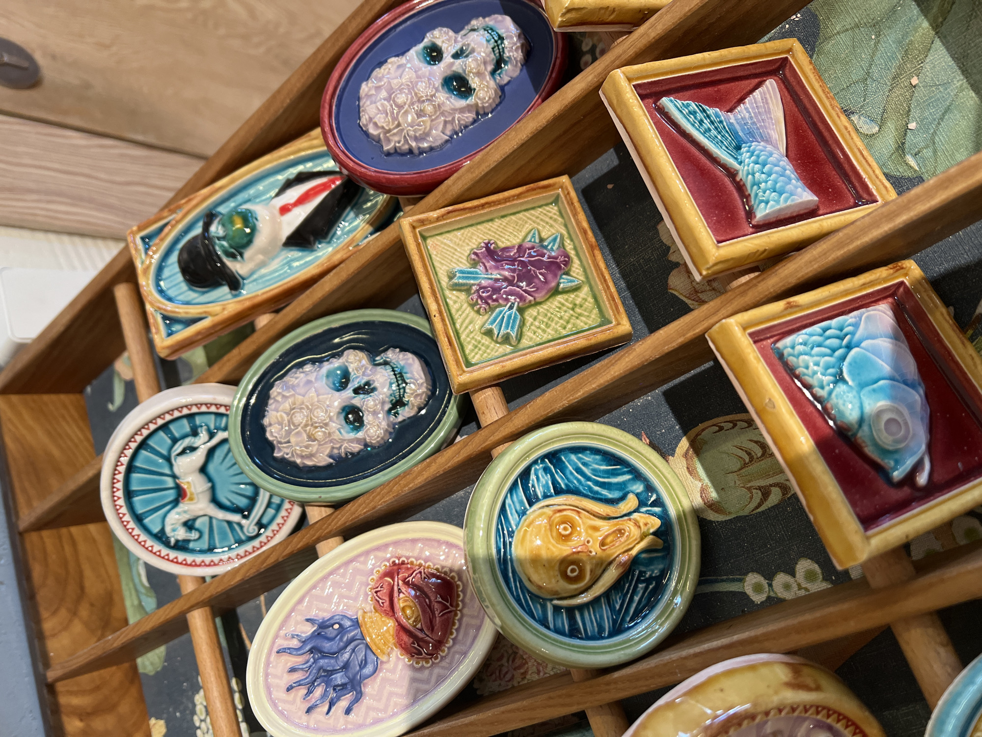 The image shows a wooden display shelf holding a collection of ceramic tiles. The tiles are arranged in rows and feature various raised designs, including: a skull, a fish, a flower, a human figure, a head, and other objects. The tiles are glazed in different colors, such as blue, green, red, yellow, and white. The background appears to be a patterned fabric or wallpaper. The location is not discernible as Moscow, Russia based solely on the visual information provided in the image.