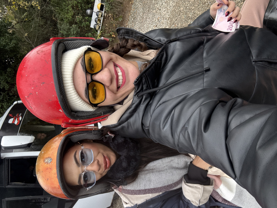 Two women are positioned in the foreground, looking directly at the camera, in an outdoor setting in Çiftlikköy, Turkey. The woman on the left wears an orange full-face helmet, clear-framed sunglasses with blue-tinted lenses, a dark fur-like collar, a striped scarf (white, pink, and beige), and a dark-colored jacket. She has dark hair and a slight smile. Behind her, a white vehicle with dark windows and a red and white logo is partially visible, with a male figure looking out from within. The woman on the right wears a red full-face helmet over a white knit beanie, black-framed sunglasses with yellow-tinted lenses, a black puffer jacket, and a beige hoodie or neck warmer. She displays a broad smile, revealing her teeth, and has dark hair in a visible braid on her right side. Her left hand, adorned with dark red, glossy, almond-shaped fingernails, holds two 200 Turkish Lira banknotes. In the background, green foliage is visible, along with several other helmets (black, yellow, white, grey) resting on a low ledge or wall. The ground appears to be a gravel or dirt surface. The scene suggests a recreational activity involving helmets, possibly ATV riding.