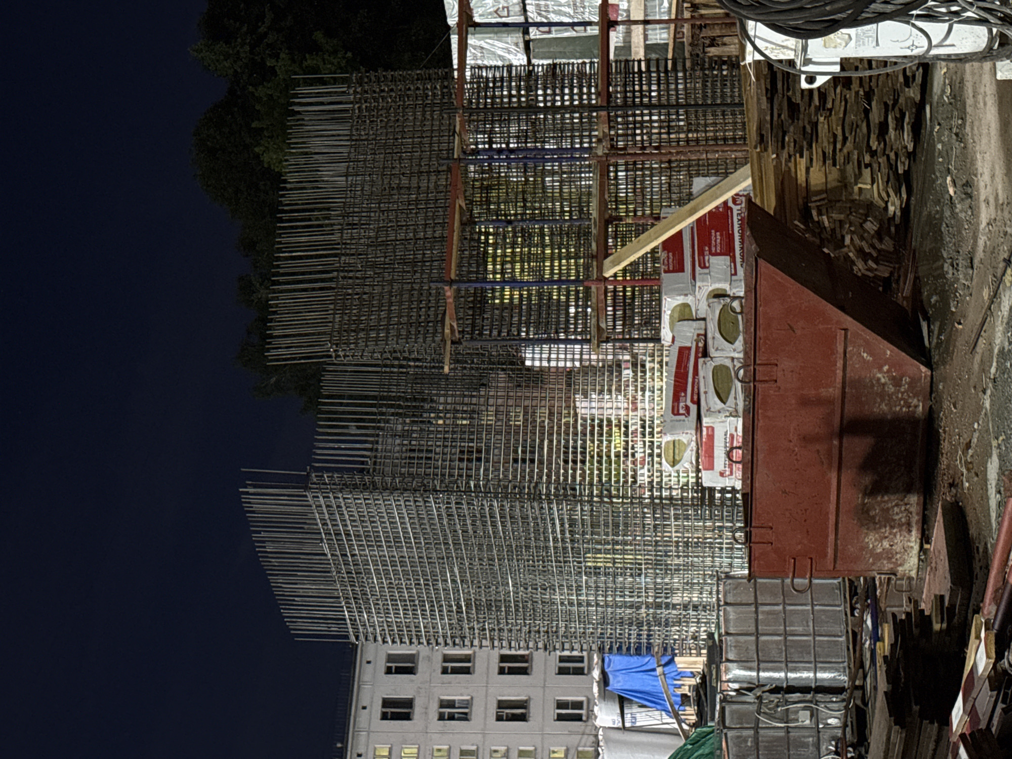 The image depicts a nighttime construction site in Moscow, Russia, focusing on the skeletal framework of a building under construction. Dominating the scene are towering structures of rebar, forming the concrete reinforcement for what appears to be a multi-story building. To the left, a light-colored apartment building with multiple windows is visible, illuminated from within. In the foreground, a large, red metal skip is positioned, along with stacks of lumber and some rolls of material covered in plastic sheeting. Several pallets of insulation are stacked near the rebar structures. The overall environment is dark, with artificial lighting from the apartment building and possibly from construction lights casting a yellowish glow on some of the rebar. The ground is uneven and appears to be dirt and debris. No individuals are discernible in the frame.