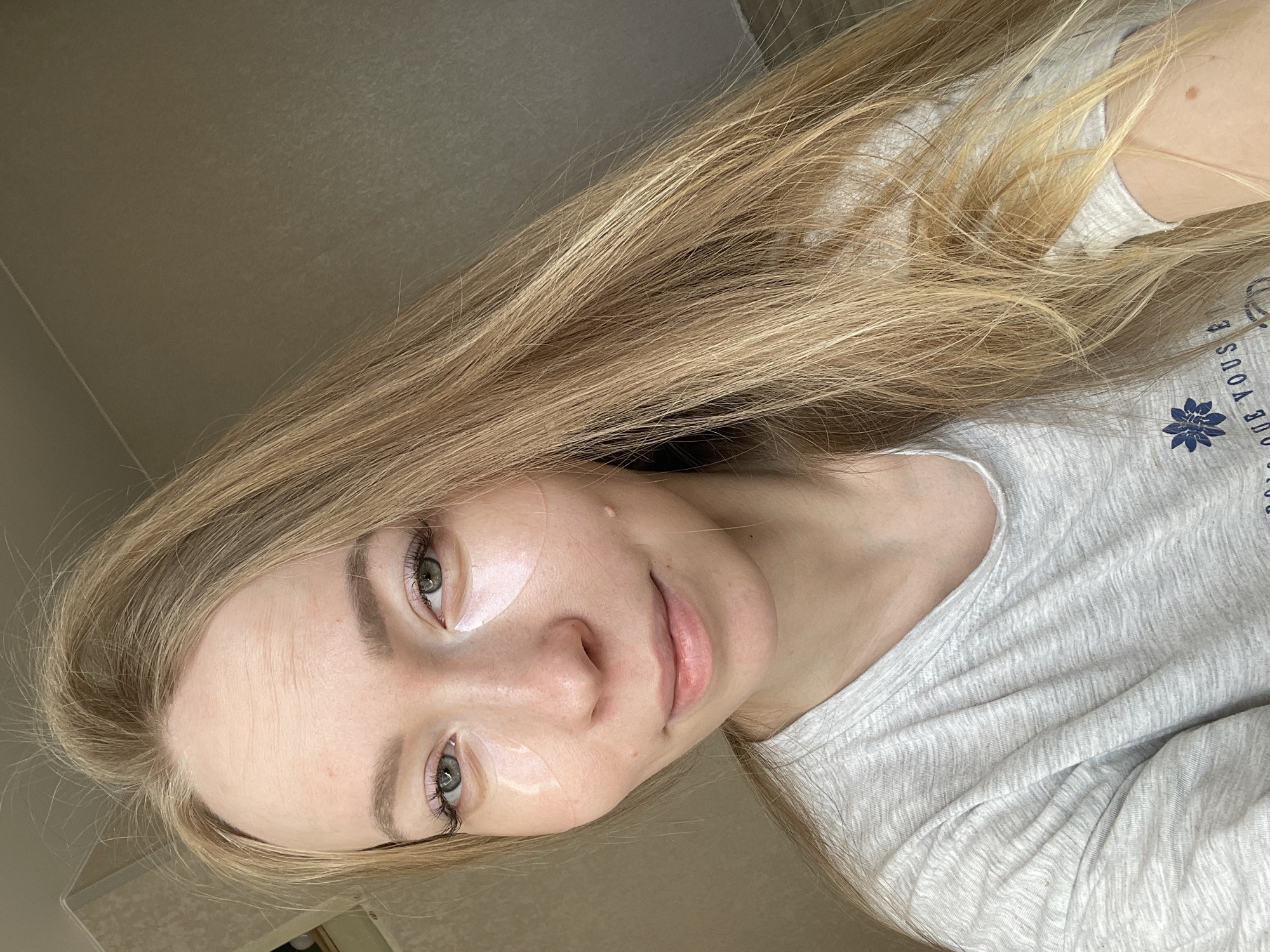A woman with long, blonde hair is pictured in a selfie. She has fair skin, green eyes, and is wearing under-eye patches. She is wearing a light-gray t-shirt with a blue floral design and lettering. The background appears to be a neutral-colored wall. There is no definitive indication of the location being Perm, Russia.