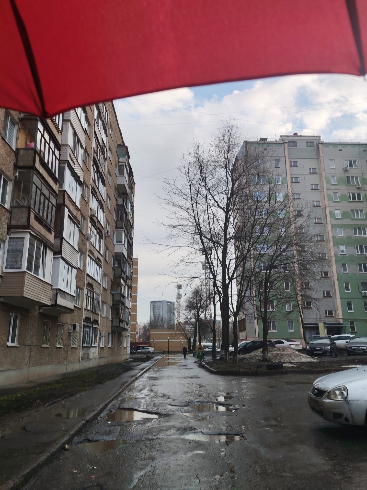 The image depicts a street scene in Izhevsk, Russia, on a rainy or post-rainy day. The view is partially framed by a red umbrella, suggesting the photographer is under cover. The street is wet and shows considerable damage with potholes filled with water.
On the left side of the street stands a tall, older apartment building with a brown facade. Several balconies are visible, some enclosed. On the right side is a newer, taller apartment building painted in grey and green tones. Bare trees stand between the street and the building.
A few cars are parked along the side of the street, with a silver car partially visible in the foreground on the right. Further down the street, a person is walking away from the camera. In the distance, a taller industrial or utility structure is visible. The sky is overcast.