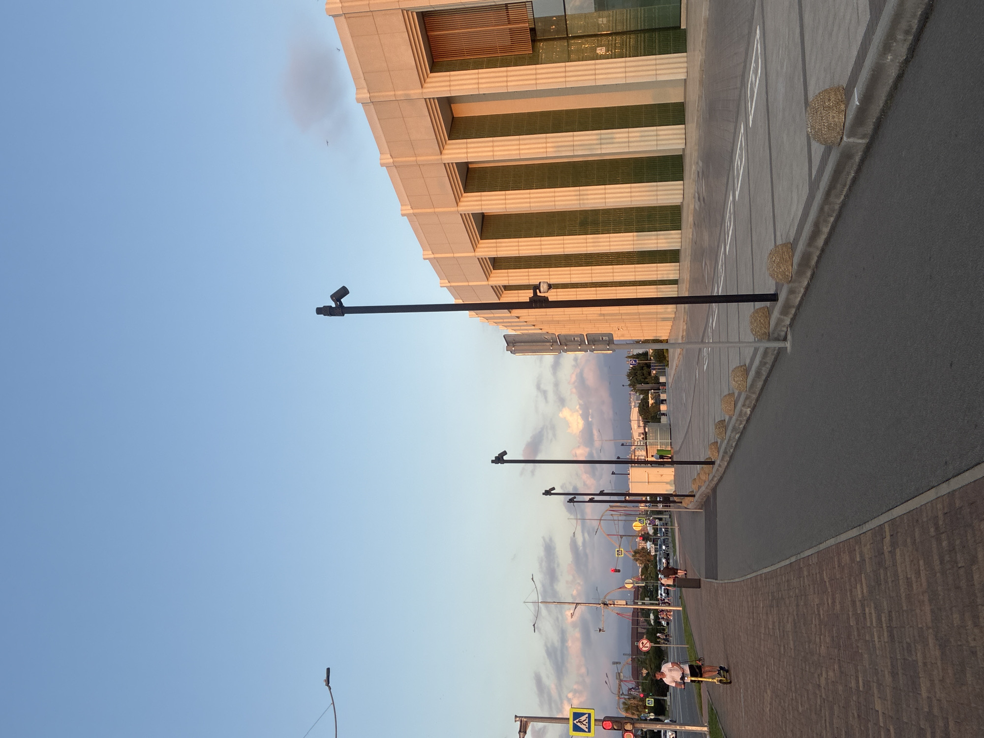 The image depicts a street scene in Sirius, Russia, during what appears to be late afternoon or early evening, with the sun casting a warm glow on the surroundings. On the right side of the frame, a large, modern building with a beige facade and vertical green panels dominates the background. The building has a contemporary architectural style, with prominent pilasters and large windows.
In the foreground, a paved pedestrian walkway runs along a brick-paved sidewalk. A person is riding a yellow electric scooter on the brick path, moving from left to right. Further down the street, a road with traffic is visible, featuring traffic lights, road signs, and several cars. Lampposts are positioned along the pedestrian walkway and the road. The sky is clear blue with some scattered, soft clouds tinged with pink and orange hues from the setting sun. The overall environment suggests an urban or suburban setting with well-maintained infrastructure.