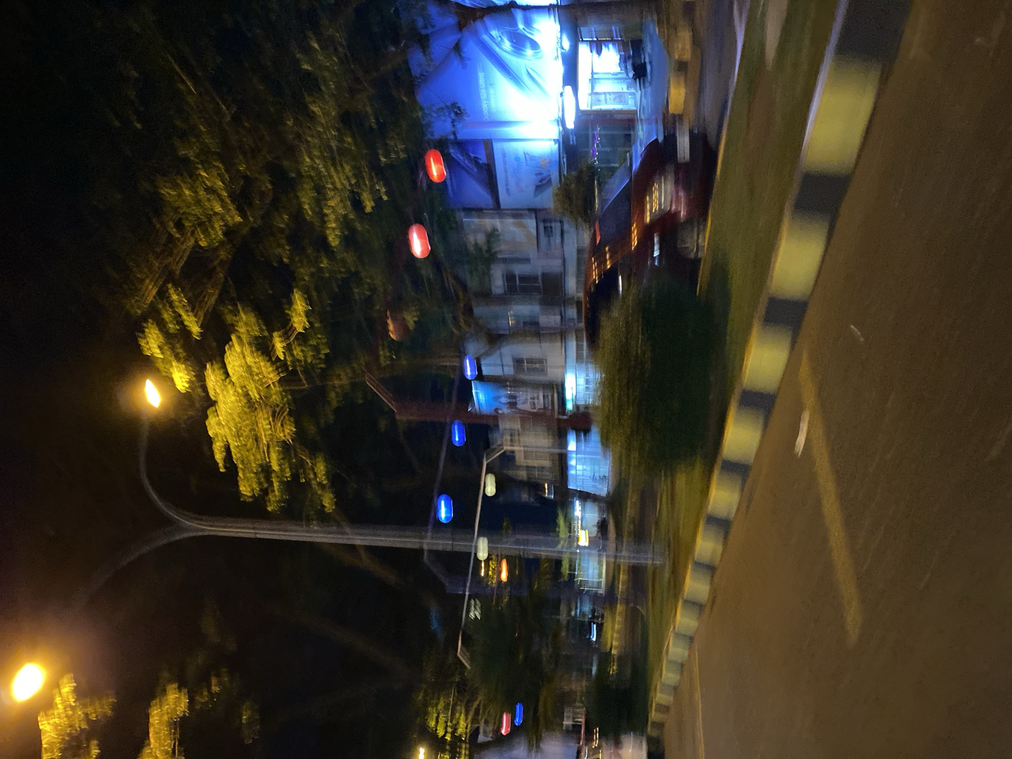 This media file captures a street scene at night in Madiun, Indonesia. The primary subjects are the street, illuminated by streetlights and decorative lanterns, and the surrounding buildings and trees. The image is characterized by motion blur, suggesting it was taken from a moving vehicle.
The scene is illuminated by artificial lights. Several lanterns, in red and blue hues, are strung across the street, adding a festive or decorative element to the urban environment. A large, brightly lit billboard with blue tones is visible in the background, displaying what appears to be a commercial advertisement. The text on the billboard is partially legible and appears to include the word "welcome" and possibly "galaxy." Adjacent to the billboard are building facades, suggesting a commercial or residential area. Lush green trees with bright yellow foliage, illuminated by streetlights, frame parts of the scene. A road with a striped median is also clearly visible, indicating a well-maintained urban thoroughfare.
The time of day is definitively night, as evidenced by the darkness of the sky and the reliance on artificial lighting. The weather appears to be clear, with no indication of rain or fog. The overall mood is energetic and vibrant, due to the colorful lights and the dynamic blur of motion. The context of Madiun, Indonesia, places the scene within a specific geographic and cultural setting. No people are clearly identifiable in the image due to the blur.