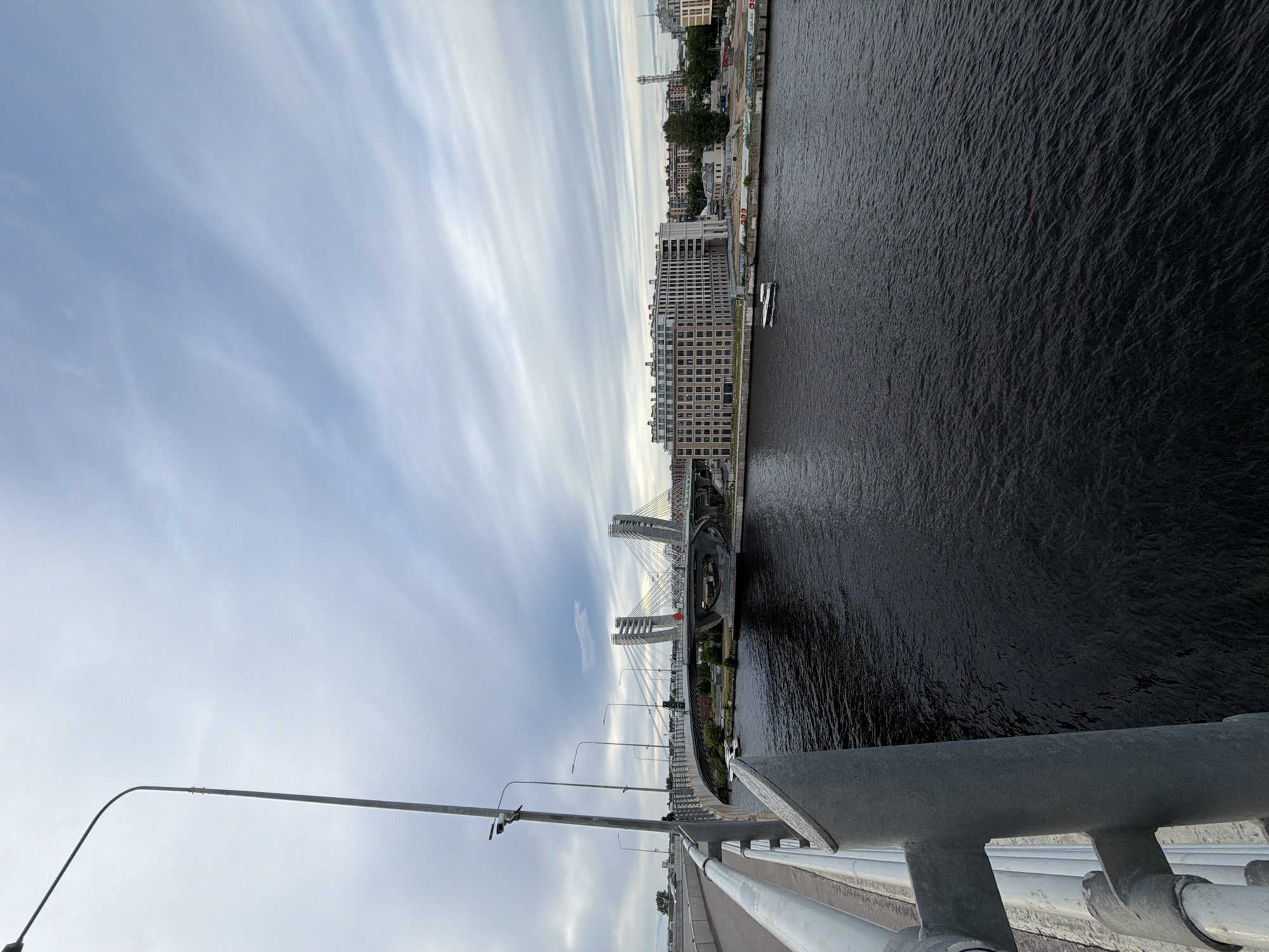 The image depicts a wide shot of a river with a modern cable-stayed bridge spanning across it. The perspective is from an elevated position, likely another bridge or structure, looking down and across the water. The river is dark and choppy, suggesting movement or wind. On the far bank, there are several buildings of varying heights and architectural styles, with a mix of residential and commercial structures. Some of these buildings are modern and tall, while others appear older and more traditional. Trees and greenery are interspersed among the buildings.
The sky is overcast with distinct streaks of clouds, indicating a potentially breezy day. The lighting suggests it is either early morning or late afternoon. The bridge itself is a prominent feature, characterized by its concrete pylons and numerous cables that support the roadway. The roadway of the bridge is visible in the foreground, with lampposts lining its edge. A metal railing runs along the edge of the viewpoint, partially obscuring the lower part of the scene. Two small white boats are visible on the water near the far bank.
There is no visible text in the image. The scene conveys a sense of urban landscape, with the bridge and city architecture dominating the view. The overall mood is somewhat somber due to the overcast sky and dark water, but also offers a sense of scale and engineering marvel in the bridge's design. The location is identified as Saint Petersburg, Russia, which is known for its numerous canals, rivers, and impressive bridges.