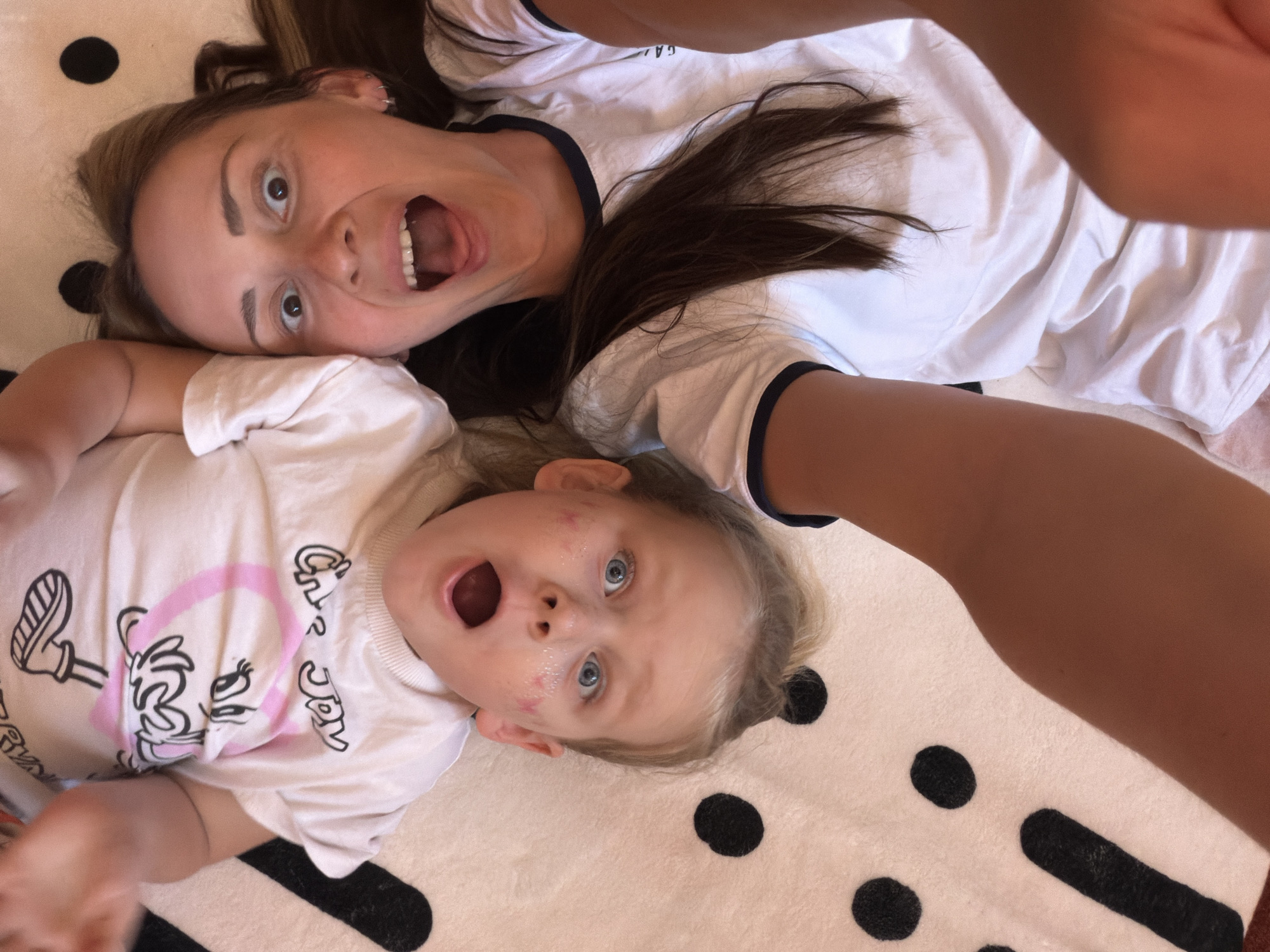 An overhead shot shows a fair-skinned woman and a fair-skinned child lying on a white rug with black circles and rectangles. The woman has brown hair, and the child has blonde hair. Both are wearing white shirts and have open mouths in apparent surprise or excitement. The photograph was taken in Khimki, Russia.