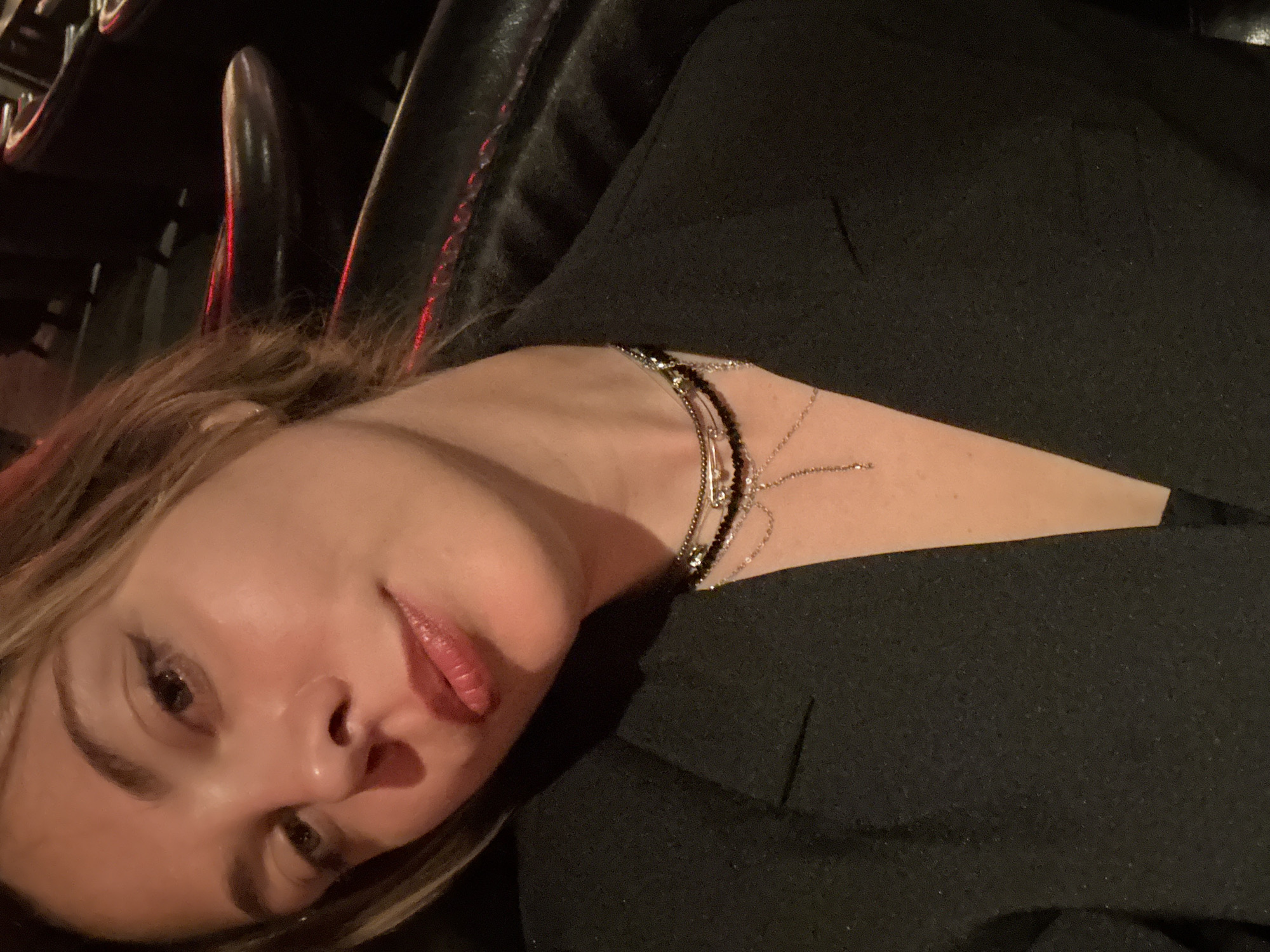 The image shows a close-up, over-the-shoulder selfie of a young woman. She has light brown hair, styled with subtle waves and bangs. Her eyes are a warm brown, and she wears dark eyeliner. Her lips are full and painted with a glossy lipstick in a pinkish-red hue. She is wearing a black blazer, and a delicate layered necklace with multiple chains and small pendants is visible around her neck. The background is dark and appears to be an indoor setting with some red lighting reflecting on what looks like leather seating, suggesting a dimly lit lounge or restaurant. There is no visual information in the image that can be used to determine the location in the city of Sinyavino, Russia.