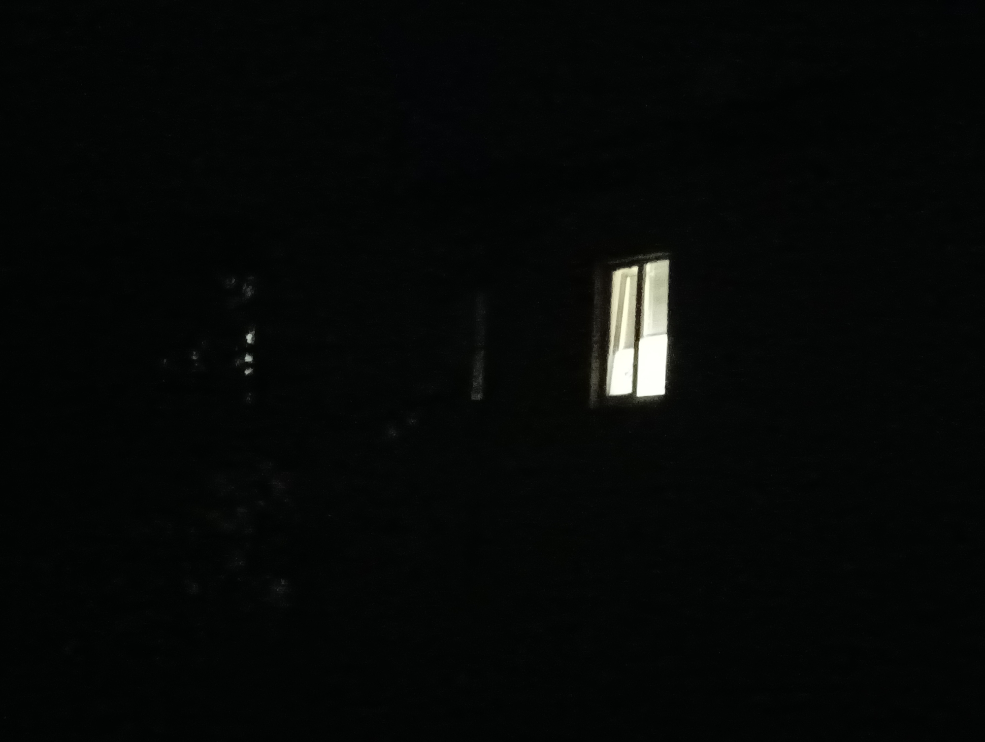 The image captures a dark, nighttime scene with a single lit window visible on a building. The window is rectangular and divided into two panes, with a warm, yellow light emanating from within. The surrounding area is almost entirely black, suggesting the photograph was taken in a rural or unlit urban setting. Faint, indistinct shapes, possibly trees or other structures, are barely discernible in the darkness to the left of the window. The overall impression is one of stillness and isolation. This scene is located in the village of Sahaidachne, Ukraine.