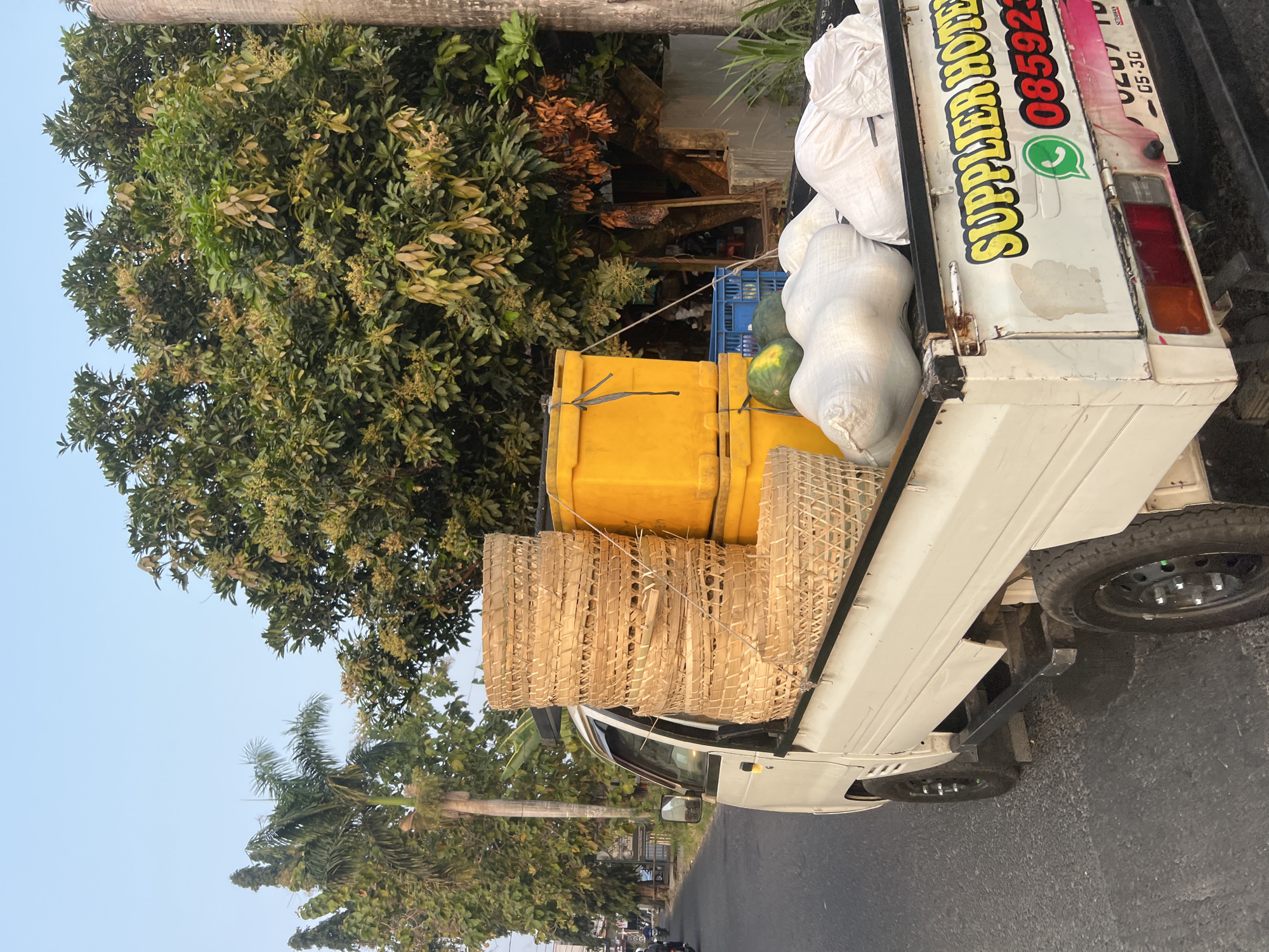 This image shows the back of a white pickup truck driving on a street in Tlogoadi, Indonesia. The truck bed is filled with various items. Stacked high are woven bamboo baskets. Next to the baskets are two yellow coolers. In front of the coolers are two watermelons. Behind the coolers, several white sacks are piled up. On the tailgate of the truck, the words "SUPPLIER HOTEL" are written in yellow with a red outline, and a green WhatsApp logo is next to a phone number that starts with "085923". Trees line the side of the road, and the sky is clear and blue, indicating daytime.