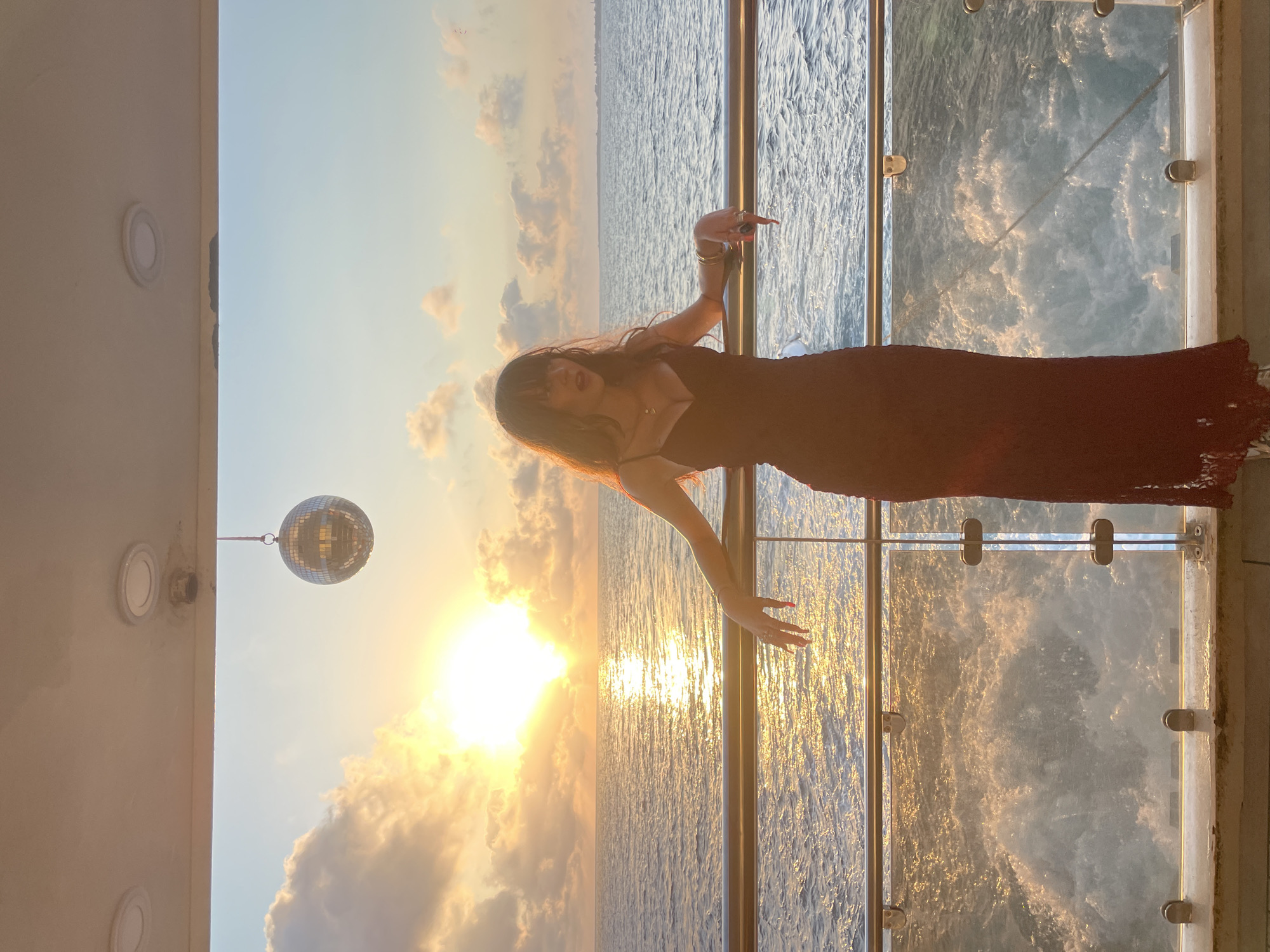 A young woman with long, wavy, dark hair and bangs stands on a boat's deck, leaning on a railing. She wears a dark, sleeveless, floor-length dress with a sweetheart neckline and lace trim at the hem. The sunset casts a warm glow over the ocean, with clouds scattered across the sky. A disco ball hangs from the ceiling above her. The boat appears to be moving, creating white wakes in the water behind it. The scene suggests a celebratory or vacation atmosphere. The location is Yeroskipou, Cyprus, specifically on a boat in the Mediterranean Sea.