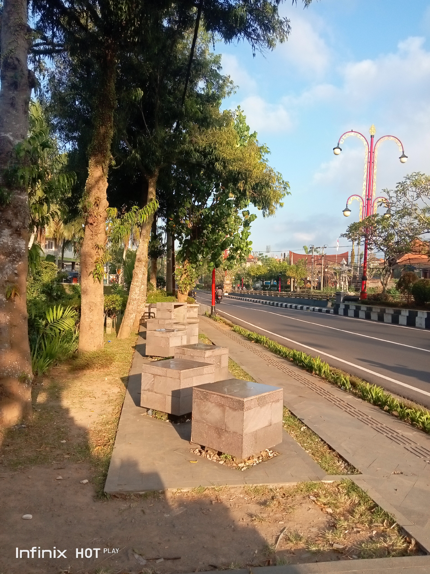 This is a photograph taken in Blahbatuh, Indonesia, showing a street scene with a sidewalk lined with trees and decorative lamp posts. In the foreground, there are several square concrete benches. The street is paved and has a white line marking the lanes. A person on a motorcycle is visible on the street. The sky is blue with some clouds, and the sunlight suggests it is either early morning or late afternoon. The overall impression is of a well-maintained urban environment with natural elements.