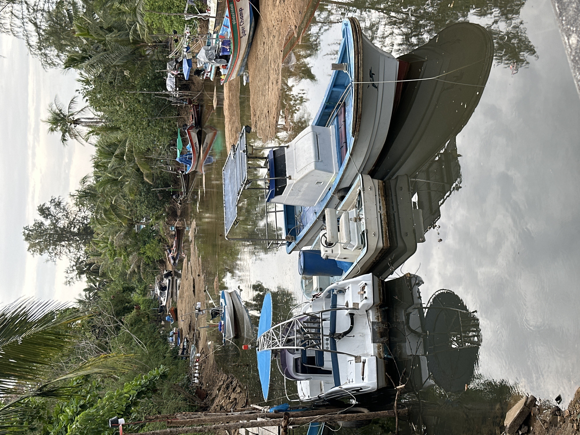 The image displays a serene waterside scene in Ko Pha Ngan, Thailand, featuring numerous small boats moored along a narrow body of water, likely a river or canal. The sky is overcast, and its cloudy reflection is clearly visible on the calm surface of the water, which also perfectly mirrors the boats and surrounding vegetation.
In the foreground and mid-ground, several white and blue boats are prominent. One white boat, with a blue trim, features a small cabin and a distinctive blue-covered structure on its stern, possibly for equipment or a small canopy. Its bow carries a small dolphin or fish emblem. Adjacent to it, another white boat is partially visible, equipped with a blue domed structure. Further down the bank, multiple other boats are lined up, some pulled partially onto the sandy or muddy shore. One boat is clearly marked with the English text "Super Champ" and "SUPER CHAMP," alongside Thai script that reads "สุเมธชัย" (Sumet Chai). Another vessel shows partial text resembling "ANPA-PHR."
The banks of the waterway are densely covered with lush tropical vegetation, including numerous tall palm trees, such as coconut palms, and various other green trees and bushes, indicating a vibrant natural environment. Wooden poles and simple structures are visible along the bank, likely used for mooring. The boats appear to be at rest, with no human activity or people visible in the frame. The scene captures a quiet moment in a local mooring area, highlighting the still waters and the natural beauty of the tropical setting under a cloudy sky.