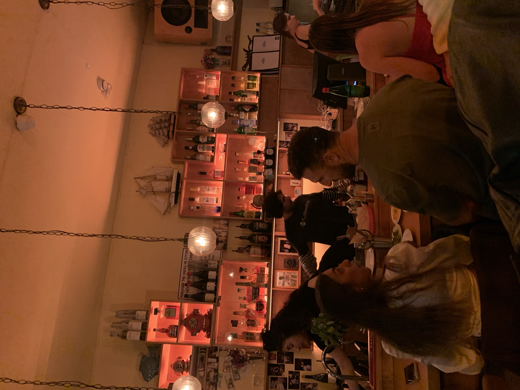 The image depicts the interior of a dimly lit bar, likely in Tel-Aviv, Israel. Several patrons stand around a bar, engaging in conversations. A variety of alcoholic beverages line shelves behind the bar. Decorations include miniature ships, a diving helmet, a fire hydrant, and numerous small photographs attached to the back wall. Hanging lights provide illumination, creating a warm ambiance.