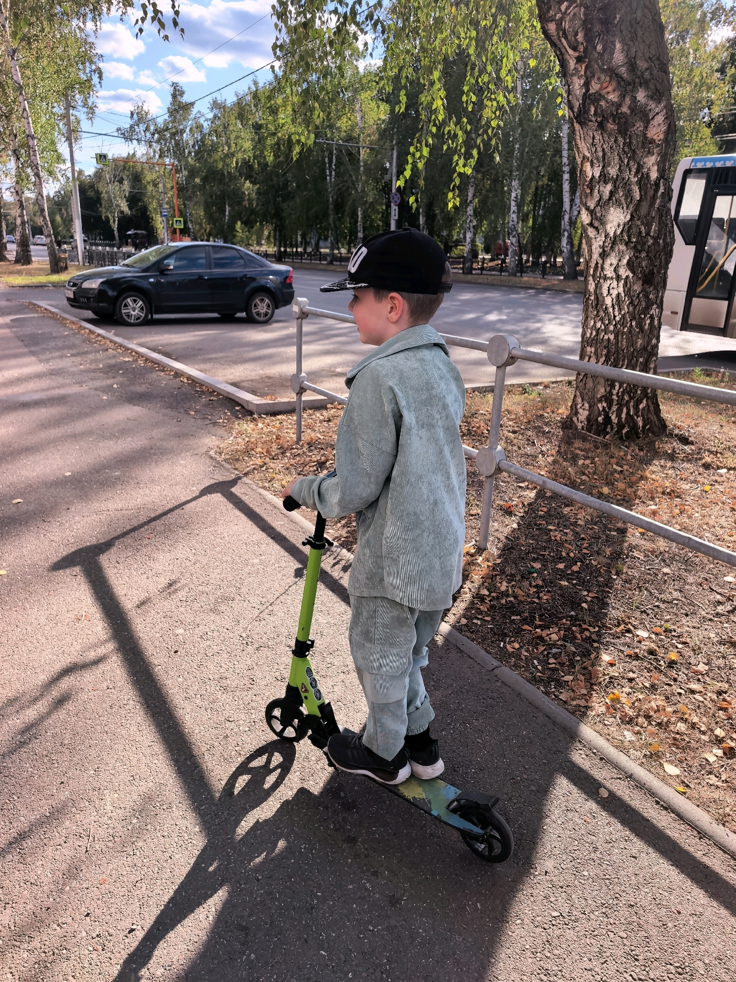 A young boy wearing a light blue corduroy suit and a black baseball cap rides a green scooter on a paved sidewalk next to a metal fence. Trees line the background, with a black car parked on the street and a white bus visible. The scene appears to be a sunny day with shadows cast on the ground. This photo was taken on Oktyabrya Avenue in Sterlitamak, Russia.