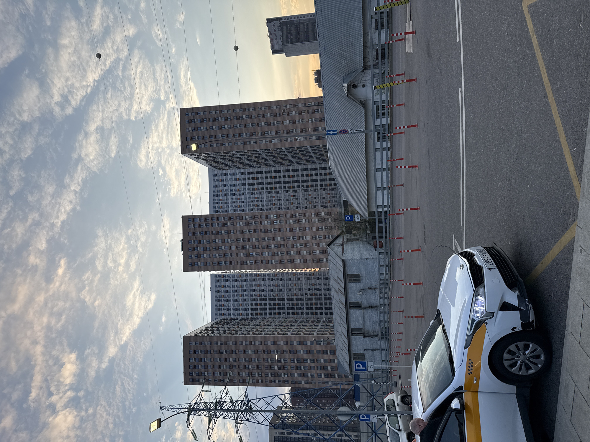 The image shows a street scene in Moscow, Russia. Several tall residential buildings are visible in the background. An overhead powerline tower is in the foreground. A white taxi with a yellow stripe is parked along the curb. Parking signs are visible. The sky is cloudy.