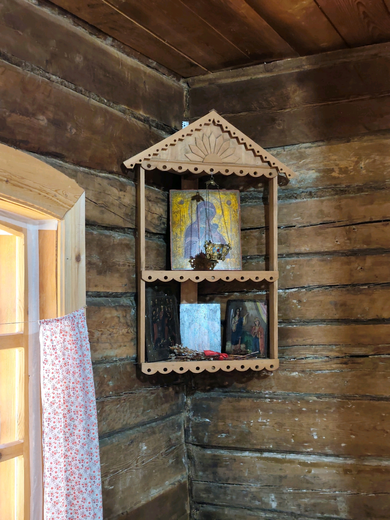 In a room constructed of dark, weathered logs, an orthodox icon shelf is mounted in the corner. The shelf is made of light-colored wood and features decorative cutouts along its edges and a peaked roof. On the top shelf, a small icon of the Virgin Mary hangs from a chain, with a brass censer suspended below it. The second shelf holds three small icons; the one on the left is mostly obscured by a blue, textured surface, while the middle icon depicts a scene with figures. To the right of this is another icon showing two figures. At the base of the shelf, a small bundle of dried branches tied with a red ribbon is visible. To the left of the icon shelf, a window with wooden framing is partly open, revealing a white curtain with a red floral pattern. The overall environment suggests a rustic interior, possibly a church or a traditional dwelling in the village of Тальцы (Irkutsk Oblast), Russia.