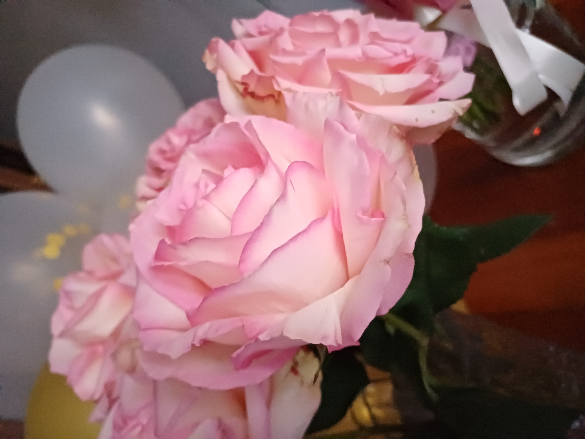 A close-up, medium shot captures a bouquet of pale pink roses with darker pink edges on their petals. The roses are arranged in the foreground, filling the majority of the frame. To the left and slightly out of focus, a light blue balloon is visible, and beneath it, a yellow balloon. In the background, a clear glass vase with a white ribbon tied around its neck is partially visible, containing more roses or greenery. The setting appears to be indoors, possibly a decorated space for a celebration or event given the presence of balloons. The lighting suggests it is daytime, but the exact time and weather conditions are not discernible. The overall mood is soft and celebratory. There is no visible text in the image.