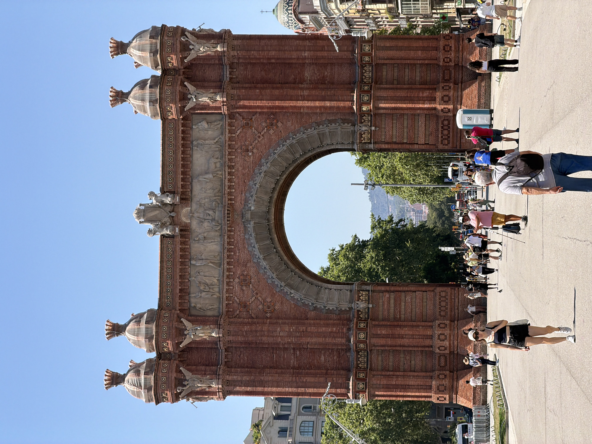 This is a full-shot, vertical, eye-level outdoor photograph taken on a sunny day in Barcelona, Spain, at the Arc de Triomf. The foreground is dominated by a paved street with scattered pedestrians. A woman in black shorts, a black tank top, and white sneakers walks towards the camera, carrying a white tote bag. Behind her, an older man with grey hair, wearing a grey polo shirt and blue jeans, walks away from the camera with a black backpack. Other people are seen walking in various directions on the street, some in groups and others individually.
The Arc de Triomf, a grand brick monument, is the central focus of the image, with its large arched opening revealing a street lined with trees and buildings beyond. The arch is adorned with sculptures and decorative brickwork. To the left of the arch, a building with a white facade and multiple windows is visible. To the right, the corner of a building with a domed roof and intricate facade is seen. The background shows a clear blue sky and a distant hilly landscape. The overall scene depicts a popular tourist area with people enjoying a clear day.