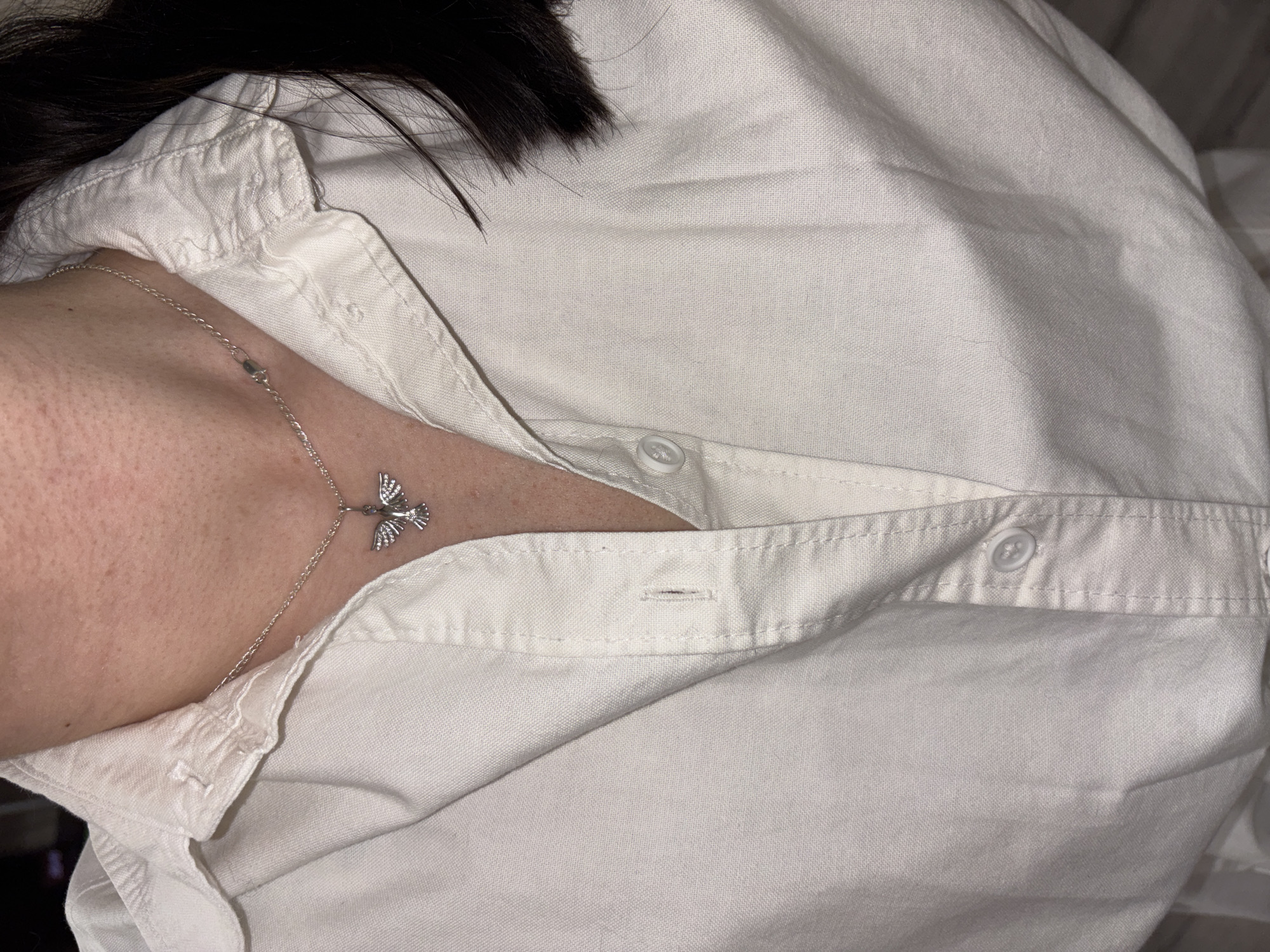 A close-up shot shows a person wearing a white button-down shirt and a silver necklace with a bird pendant. The shirt is partially unbuttoned, revealing a portion of the person's skin and the necklace. Some dark hair is visible at the top of the frame. The background is out of focus. No evidence suggests that this photo was taken in Perm, Russia.