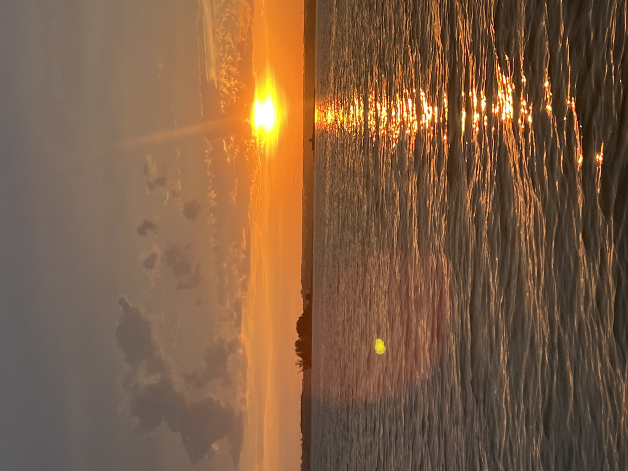 A body of water reflects the bright orange glow of a sunset. The sun, a brilliant white orb, is low on the horizon, casting a shimmering path across the water. Silhouetted against the vibrant sky, a strip of land can be seen with trees and structures on it. In the distance, a ship with cranes is visible. The sky above is a gradient of warm orange and cooler grays, with scattered clouds. A lens flare creates a bright yellow circular artifact on the water in the foreground. The overall scene captures a tranquil moment at sunset over a body of water, possibly the Black Sea near Beregovoe, Russia.