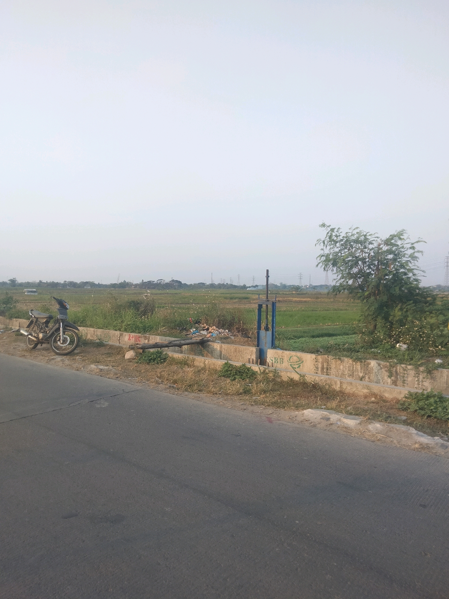 A motorcycle is parked on the side of a paved road in Suka Sari, Indonesia, next to a grassy area with a concrete channel. A metal gate, likely a water control mechanism, stands in the channel. Beyond the channel, there is a green field, possibly a rice paddy, with scattered debris. In the background, power lines and trees are visible under a hazy sky. The scene appears to be a rural or semi-rural area.