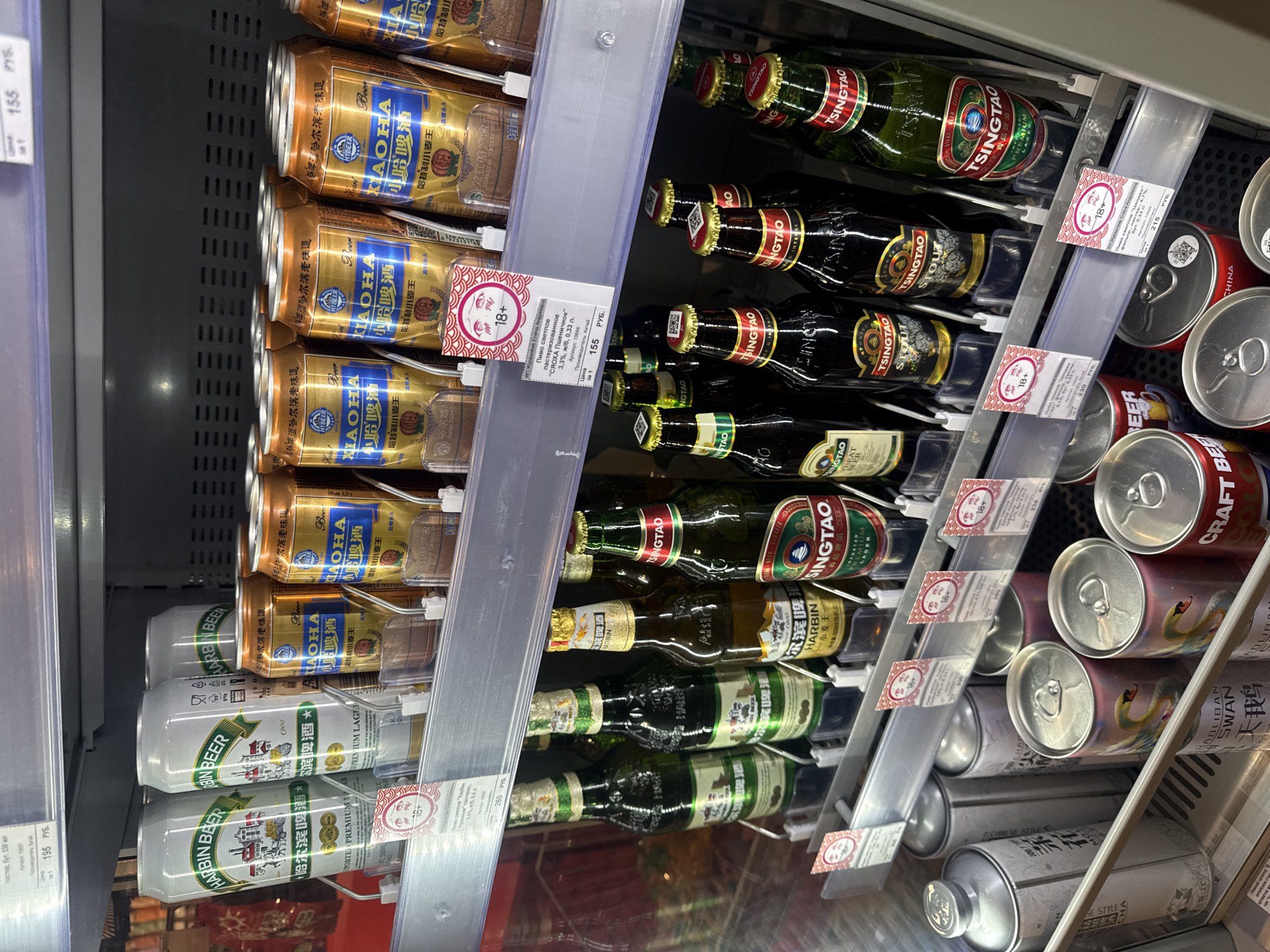 The image shows a refrigerated shelf in a store, likely in Saint Petersburg, Russia, stocked with various beers. On the left, several cans of Harbin Beer and Xiao Ha Beer are stacked. In the center, there are bottles of Tsingtao beer, including lager, stout, and wheat beer varieties. On the right, there are cans of Craft Beer and Mad Liban Swan Beer. Price tags with Russian text indicating the price per item and age restriction warnings (18+) are attached to the shelves in front of the beer selections. The brands suggest a selection of both local Russian and imported Chinese beers.