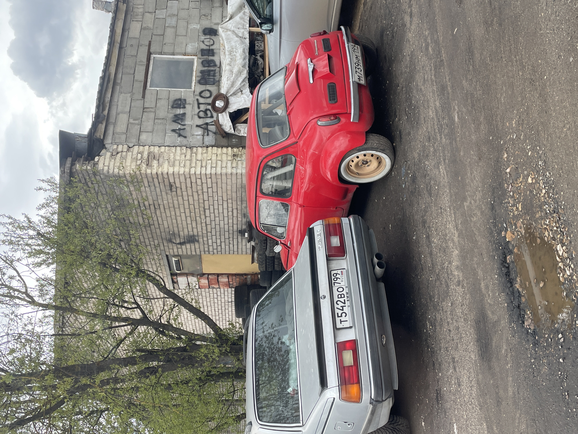 The image shows a street scene in Mar’ino, Russia. A red compact car and a silver BMW 7-Series are parked next to each other. Behind the cars is a weathered, two-story brick building with graffiti and a pile of tires. A tree with fresh green leaves is visible on the left. The street is paved with asphalt and shows signs of disrepair, including potholes. The sky is overcast. A sign on the back of the silver car reads: 'T542BO 799'. The license plate on the red car appears to read: 'M739HM 199'.