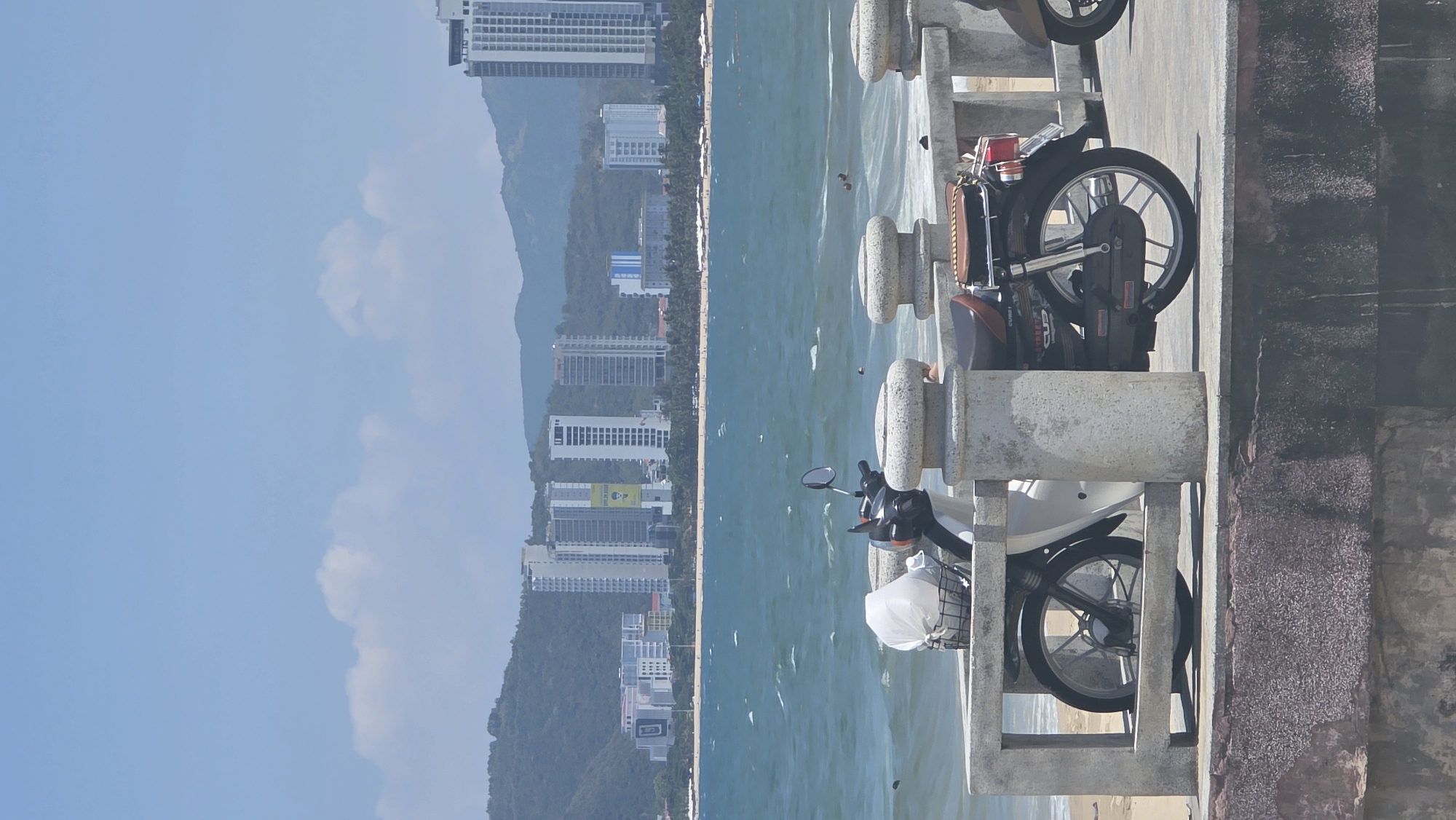 The image shows a coastline scene in Nha Trang Ward, Vietnam, with a pier in the foreground. Two motorbikes are parked on the pier. The backdrop features a beach, the ocean, and a cityscape with numerous high-rise buildings nestled against a mountain range under a partly cloudy sky. People can be seen in the water.