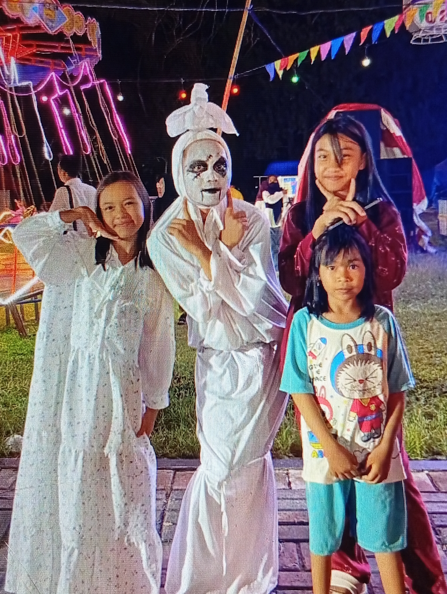 Four people are posing for a photo in front of a brightly lit amusement ride at night. The person in the center is wearing a white costume with face paint resembling a ghost or a mime, complete with dark eye makeup and a stark white face. To the left, a young person in a long, white, tiered dress smiles and poses with a hand near their face. To the right of the person in the costume, an older young person in a dark red outfit with long dark hair smiles and makes a peace sign with their fingers. In front of them and slightly to the right, a younger child with short dark hair wears a colorful t-shirt with a cartoon cat and blue shorts. In the background, colorful lights from an amusement ride and string lights are visible, along with a dark sky. The ground appears to be paved, with some grass visible to the left. The overall scene suggests a festive or event atmosphere at a fairground or amusement park.