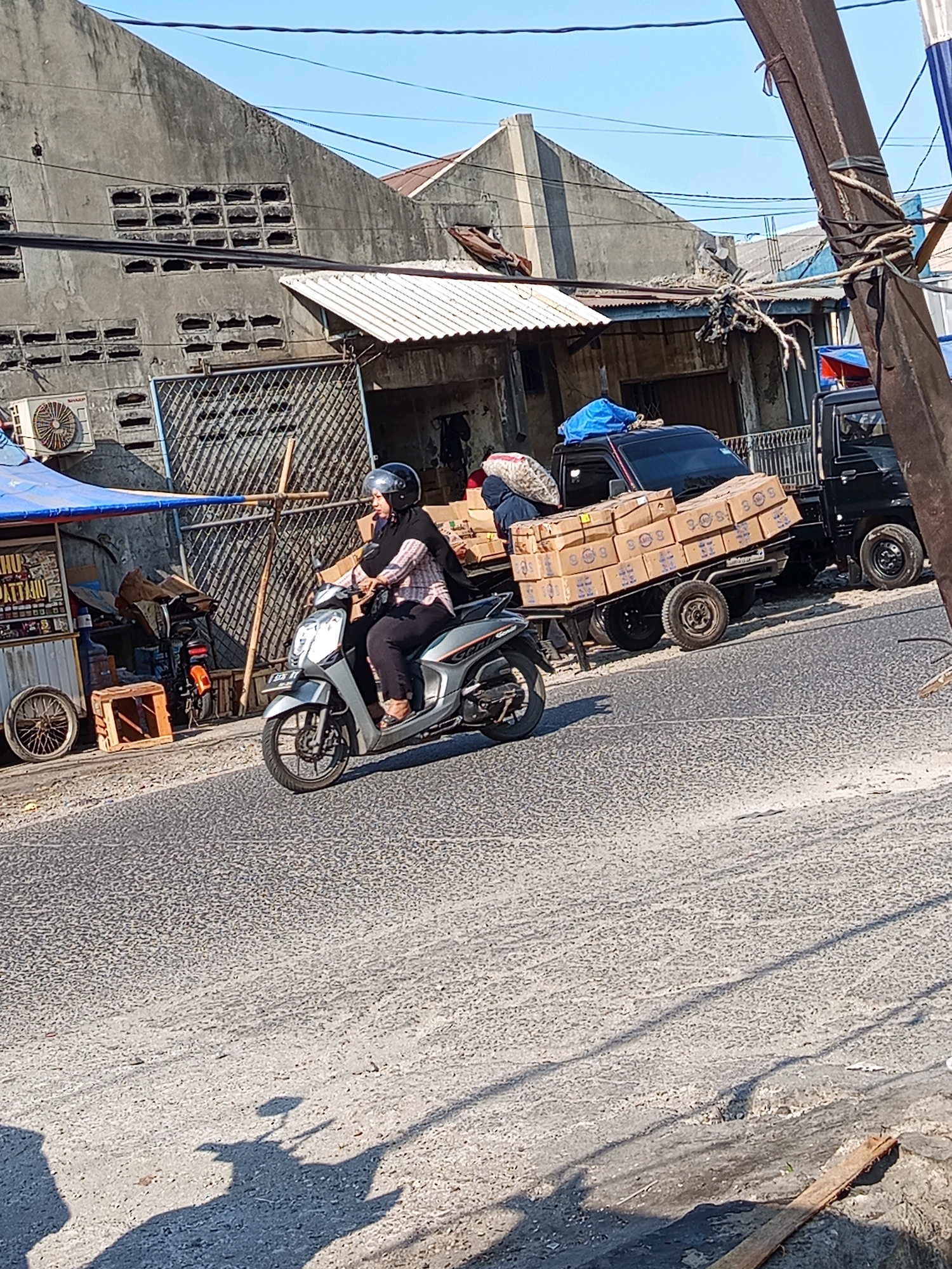 In Karawang, Indonesia, under a clear blue sky, a woman wearing a black hijab and a patterned shirt rides a silver scooter down a paved road. She is carrying a black bag and wearing a black helmet. Behind her, a black pickup truck tows a trailer filled with stacked cardboard boxes, some marked with red lettering. The truck has a blue tarp covering items in its bed. To the left of the road, a small blue-topped stall displays items and has a bicycle wheel attached. In the background, buildings with corrugated roofs and concrete walls are visible. The road surface is rough and grey, with shadows of vehicles and a utility pole casting onto it.