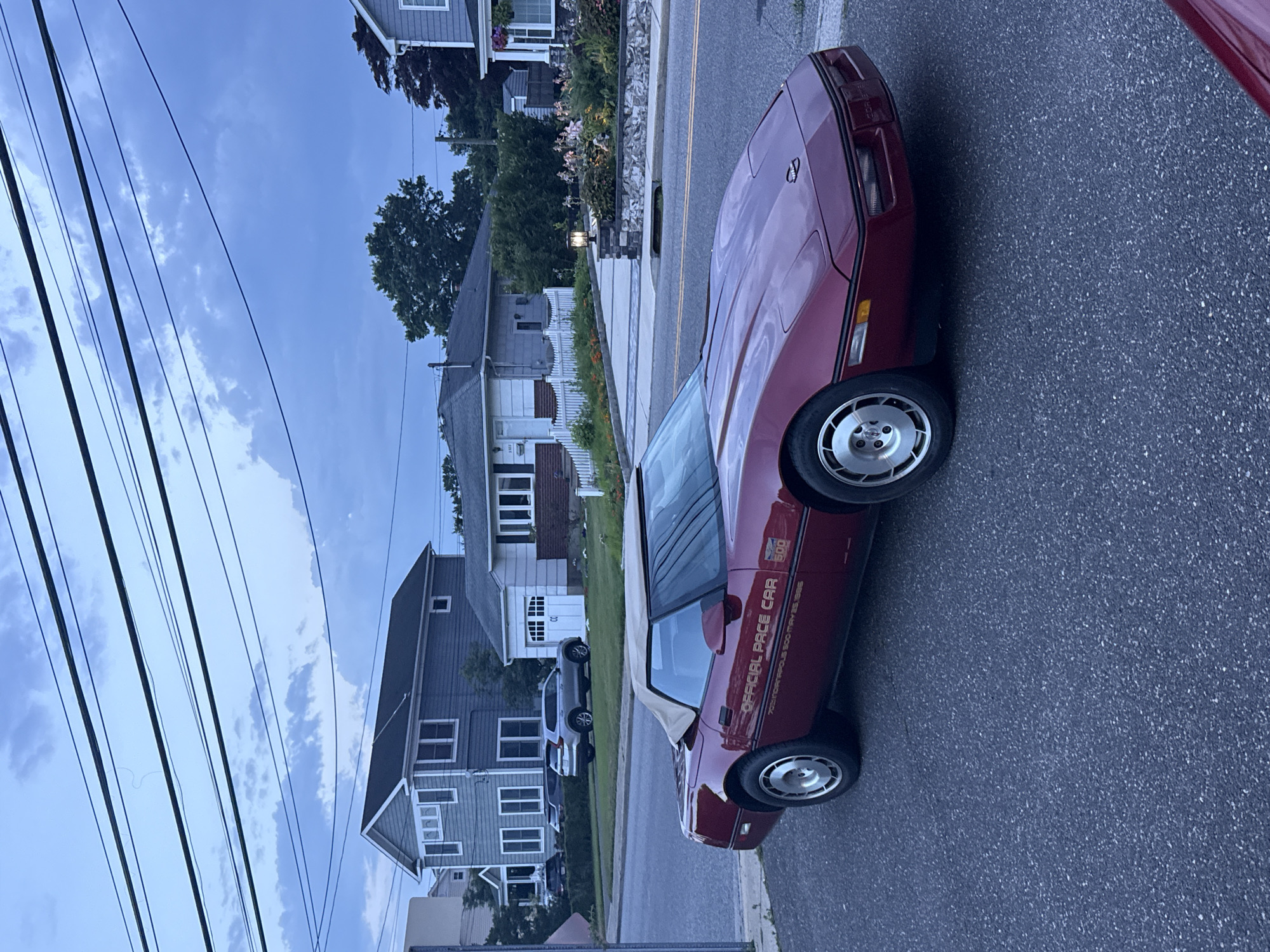 A maroon convertible Chevrolet Corvette, identified as the "OFFICIAL PACE CAR 70TH INDIANAPOLIS 500 MAY 25, 1986," is parked on an asphalt street in Bellmore, United States of America. The car is facing right, with its driver's side partially visible to the viewer. In the background, a two-story grey house with white trim and windows is visible, along with a smaller white house with a garage door and white picket fence. Trees and power lines are also present in the scene, under a cloudy sky. The sky is a deep blue with scattered white clouds. The overall lighting suggests it is late afternoon or early evening.