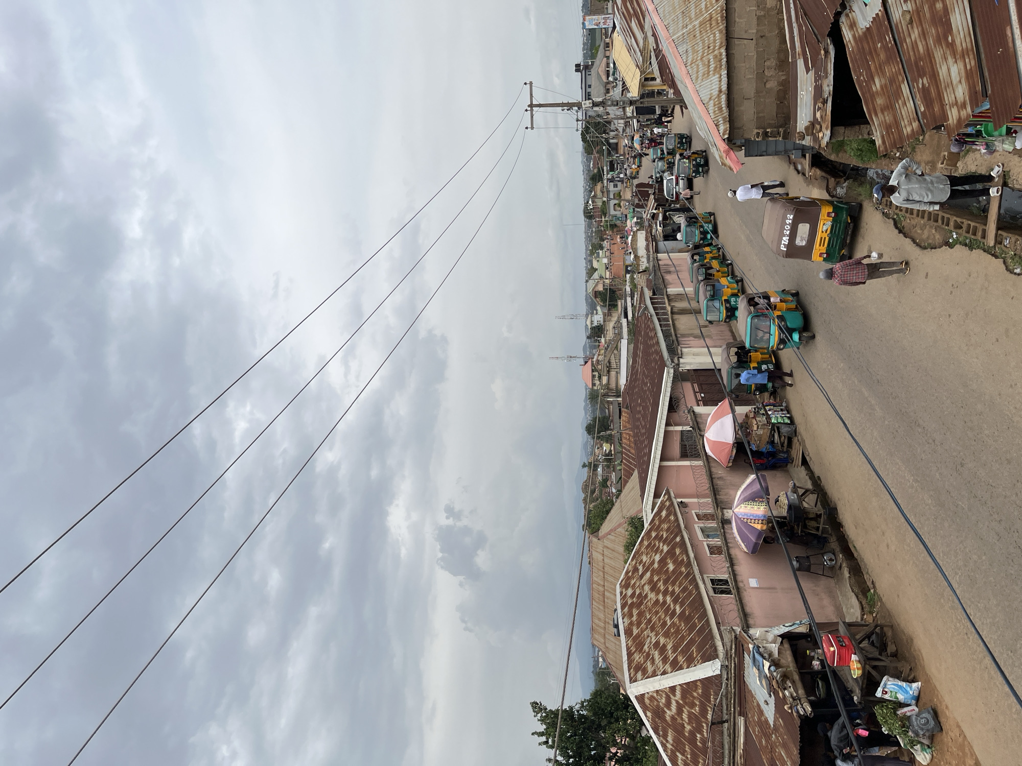This image captures an elevated view of a bustling street scene in Gura Top, Nigeria, on an overcast day. The street, which appears to be unpaved in parts, is lined with numerous buildings and a prominent row of parked tricycles (tuk-tuks).
In the foreground and along the right side of the road, a long line of green and yellow tricycles is visible, possibly waiting for passengers or parked. One tricycle clearly displays the text "PTA 2042" on its rear. Several people are scattered along the roadside; some are walking, while others are standing or congregating, possibly engaged in roadside commerce. On the left side of the street, two large, colorful umbrellas – one red and white, and another purple – provide shade for vendors or market stalls, with various goods and structures visible beneath them.
The buildings lining both sides of the street feature a mix of painted walls, often in pastel or muted tones, and many have rusted corrugated metal roofs, indicating age and exposure to the elements. Utility poles and a network of overhead power lines crisscross the sky, particularly on the left side of the frame, contributing to the urban infrastructure. Open gutters or drains run parallel to the road in several sections. The background reveals more residential or commercial structures stretching into the distance, with faint outlines of hills or mountains under a heavily clouded sky. The scene depicts a lively local thoroughfare during daytime.