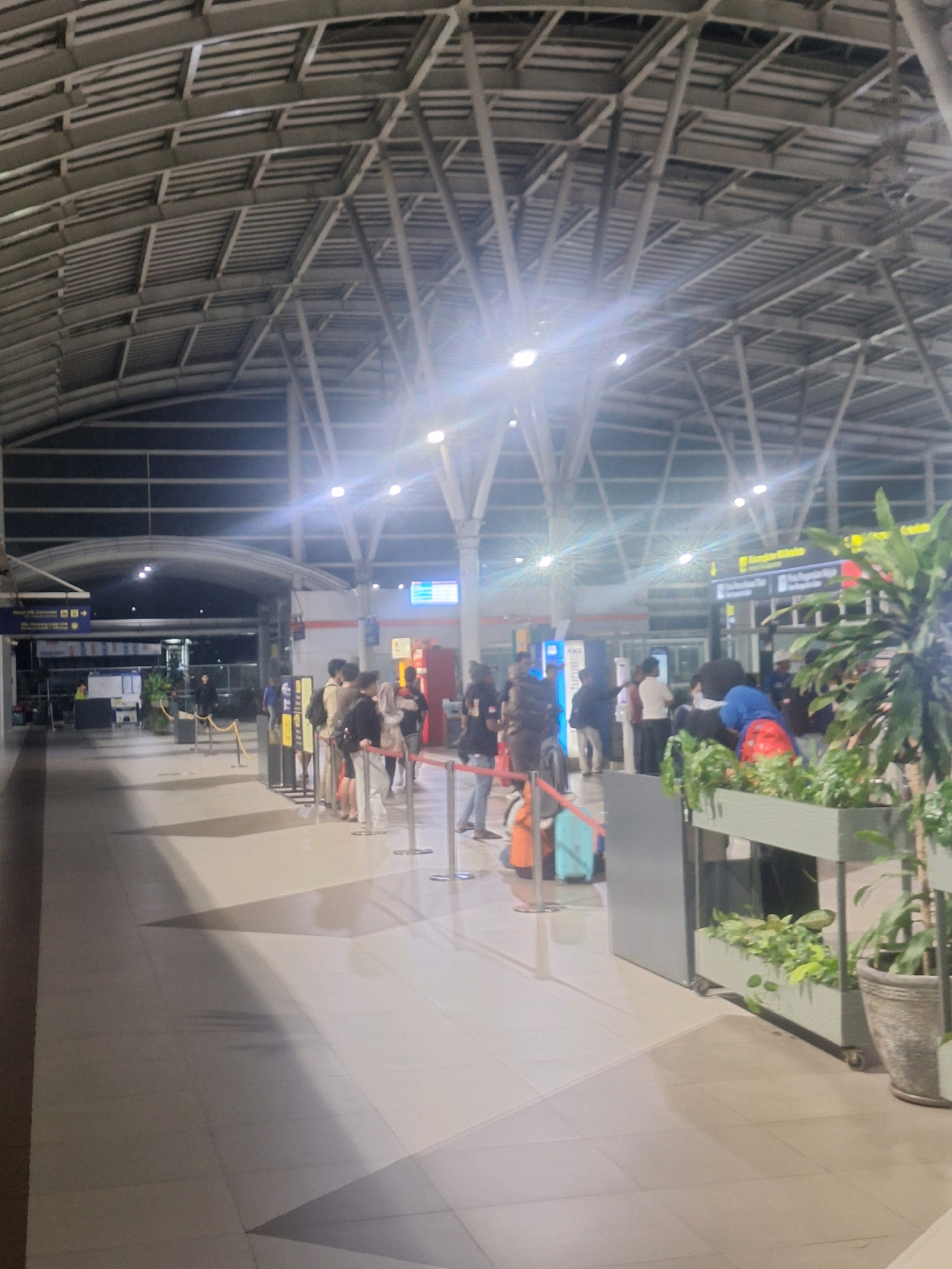 The image depicts the interior of a train station in Bekasi, Indonesia, likely during the evening or night, given the lighting conditions and the illuminated signs. The architecture features a high, arched ceiling constructed of metal beams and panels, with fluorescent lighting fixtures casting a bright glow and lens flares.
In the foreground, a tiled floor stretches across the spacious hall. Several groups of people are visible, appearing to be passengers. Some are standing in lines, separated by stanchions and red ropes, suggesting queue areas for ticketing, boarding, or information. Luggage, including suitcases and backpacks, is present with the travelers. A large potted plant in a grey planter is situated on the right side of the frame.
Further into the station, more people are gathered, and various signs are mounted on the walls and pillars, some of which are illuminated. These signs appear to provide directional information or service details, common in transportation hubs. The overall atmosphere is one of moderate activity, typical of a public transit facility.