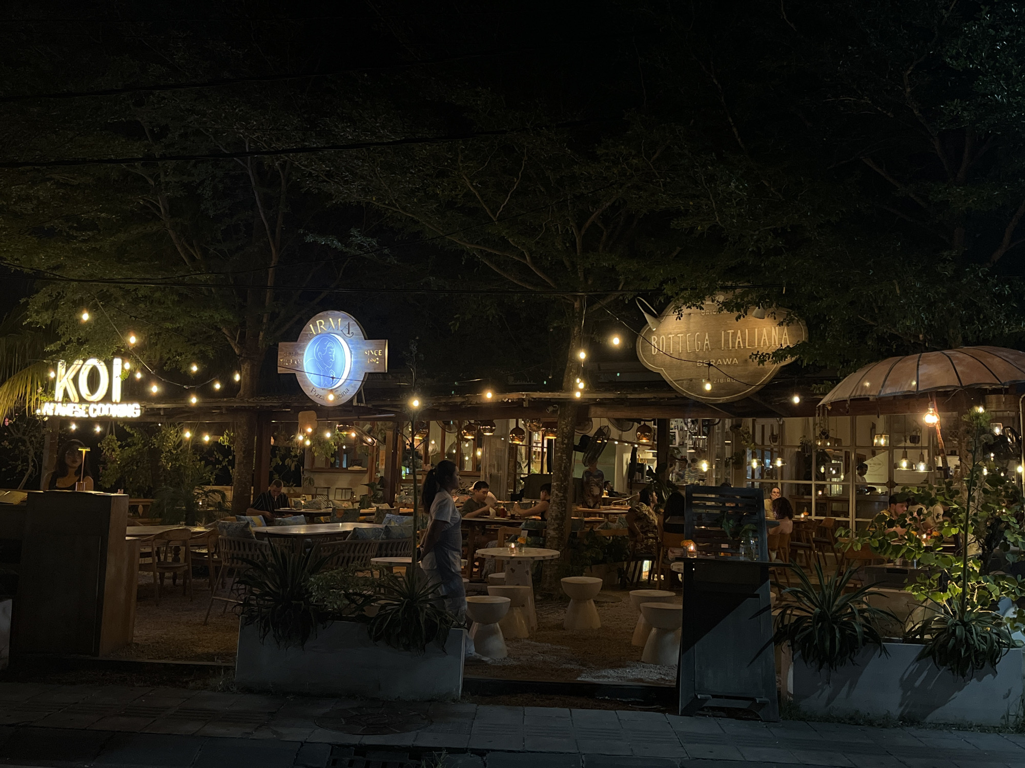The image shows a lively outdoor restaurant scene at night in North Kuta, Indonesia. The establishment appears to be named "KOI Japanese Cooking" on the left, indicated by a neon sign. To the right, another sign reads "BOTTEGA ITALIANA BERAWA," suggesting a split or adjacent establishment with Italian cuisine. String lights and numerous pendant lamps illuminate the outdoor seating area, creating a warm and inviting ambiance.
Several patrons are seated at tables, some engaged in conversation, others reading or dining. A server, wearing a light-colored top and apron, stands near the foreground, observing the diners. The restaurant features a mix of seating, including wooden chairs and tables, as well as more modern, stool-like seating. Lush greenery is incorporated into the restaurant's design, with plants in planters and trees providing a natural backdrop. The overall impression is that of a popular dining spot attracting both locals and tourists. The street in front of the restaurant is paved, and shadows are cast by the overhead lighting.