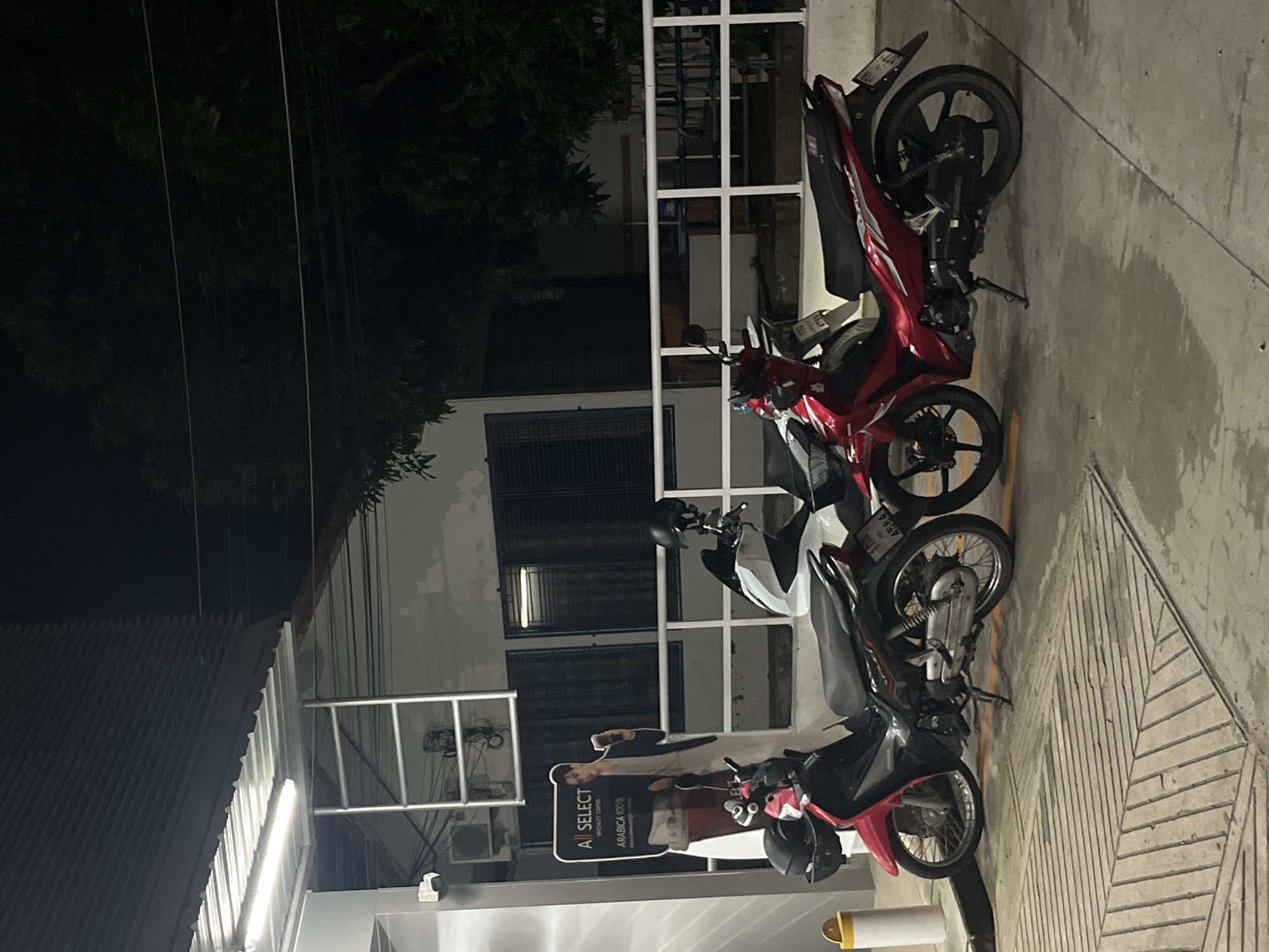 The image depicts a nighttime scene outside a building in Rawai, Thailand. In the foreground, three motorcycles are parked. The leftmost motorcycle is red and black, the middle one is white and black, and the rightmost is red. All appear to be standard street bikes. A life-sized cardboard cutout of a young man holding a coffee cup stands near the motorcycles, advertising "ALL SELECT SPECIALTY COFFEE ARABICA 100%". The building in the background has a white facade and dark-framed windows. A white metal railing runs horizontally across the middle ground. Overhead, a metal roof structure is illuminated by fluorescent lights, and trees are visible in the dark background. The ground is paved and wet in places, suggesting recent rain or cleaning.