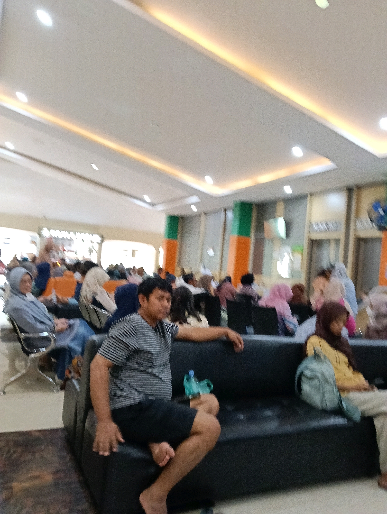 A moderately blurry, medium-shot, indoor photograph captures a waiting area with several individuals seated on a large, dark-colored sofa and individual chairs. The lighting is bright, with a lit ceiling featuring recessed lights and an illuminated cove along the edge of the ceiling. The overall impression is of a public waiting room, possibly in a medical facility or government office.
In the foreground, a man wearing a black and grey striped short-sleeved shirt and black shorts is seated on the sofa, with his legs crossed and his feet bare. His left hand rests on the sofa beside him. To his right, on the same sofa, a person wearing a yellow shirt and brown headscarf is seated. A light blue backpack rests on the sofa between them.
Further in the background, other individuals are visible sitting on chairs and the sofa, many of them wearing headscarves, indicating a likely Islamic cultural context. Some are looking towards the foreground, while others are looking away. The chairs are a mix of colors, including orange and grey. The walls have vertical accents in orange and green. The flooring appears to be light-colored tile. The scene is suggestive of a waiting room in Magelang, Indonesia, given the visual cues. The blurriness of the image prevents detailed identification of facial features or specific activities beyond sitting and waiting.