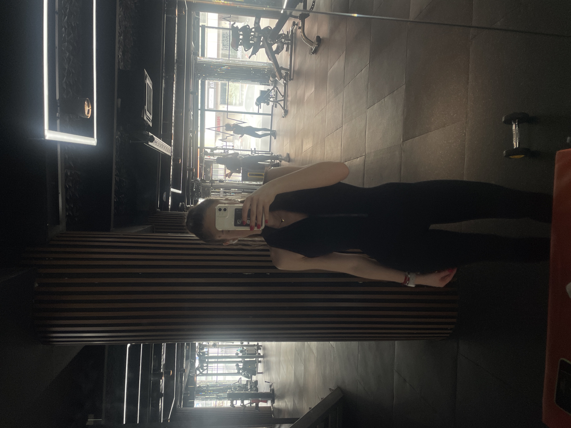 A female subject is depicted in a full-length mirror selfie taken within a modern gymnasium. The subject, centered in the frame, holds a white smartphone, partially obscuring her face, to capture her reflection. She wears a black, form-fitting, halter-neck athletic jumpsuit with a central zipper. Red nail polish is visible on her fingers, and a small gold-colored pendant hangs from her neck. A red and white bracelet adorns her left wrist. Her hair is pulled back.
The gym interior features a dark grey rubber-tiled floor and a prominent, cylindrical column clad with vertical wooden slats in varying brown tones. The dark ceiling displays multiple rectangular LED strip lights and a visible air conditioning unit.
In the background, through large windows, an urban street scene is visible. Reflections on the window glass reveal Cyrillic text, specifically "ПАРКИНГ" (PARKING), confirming the location as Sofia, Bulgaria. Two additional female individuals are visible in the gym's reflection: one, taller, in light athletic wear, stands to the left of the windows; another, in dark athletic attire, stands further right near the windows. Various gym equipment, including cable machines, weight racks, and benches, are dispersed throughout the mirrored area. A small dumbbell with a chrome handle and black, yellow-accented weights rests on the floor in the lower right foreground of the reflection. The scene is illuminated by artificial overhead lighting and natural light from the windows, producing reflections and glares on the mirror surface.