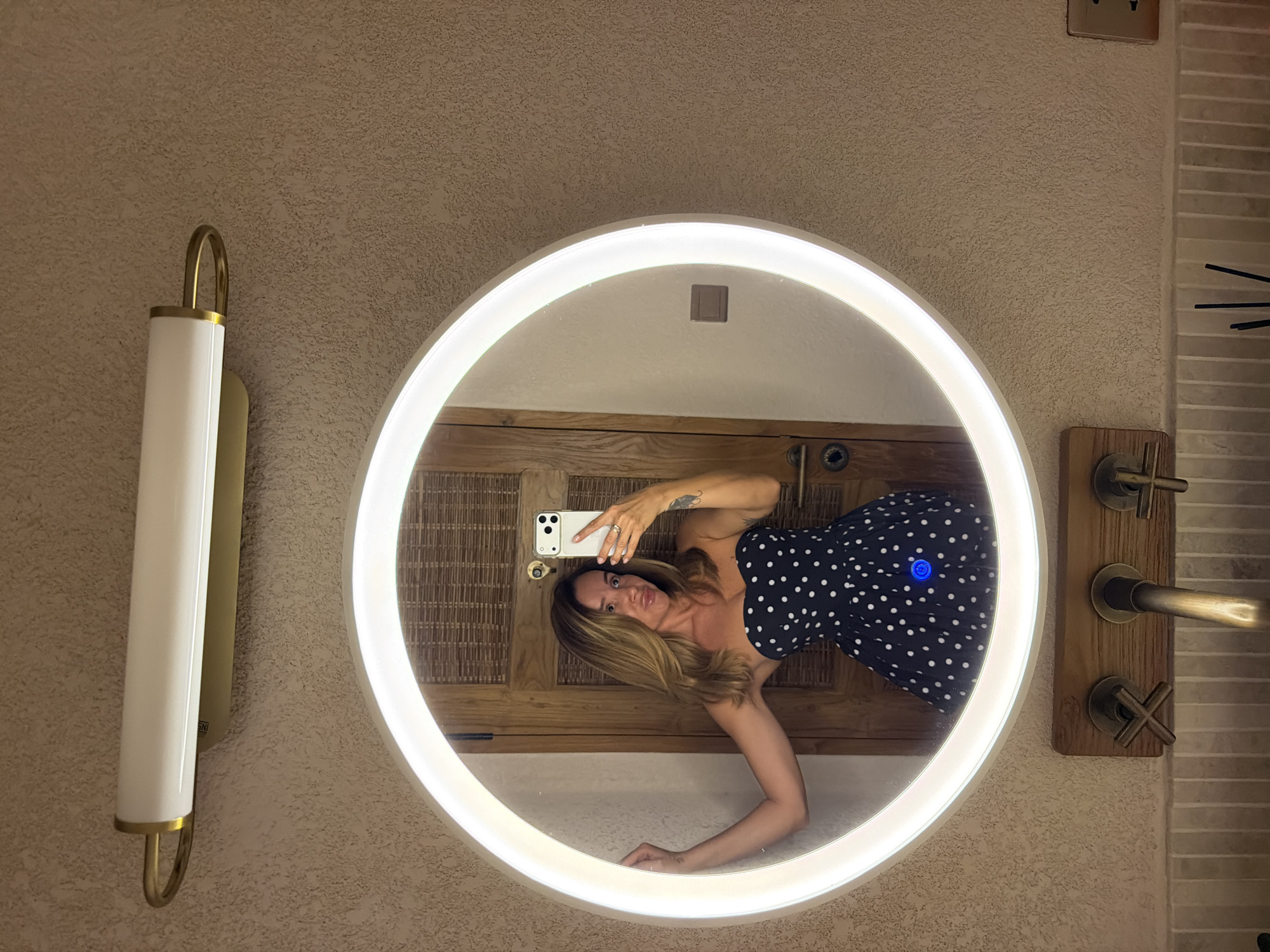 A woman is taking a mirror selfie in what appears to be a bathroom. She is wearing a strapless black dress with white polka dots. She has blonde hair and is holding a smartphone in her right hand. The mirror is circular and illuminated by an LED light. Above the mirror is a horizontal light fixture with a white shade and gold accents. Below the mirror are two vintage-style faucets and a wooden mounting. The wall behind the mirror has a textured finish. The background visible in the mirror includes a wooden door with rattan paneling. The location is identified as Mengwi, Indonesia.